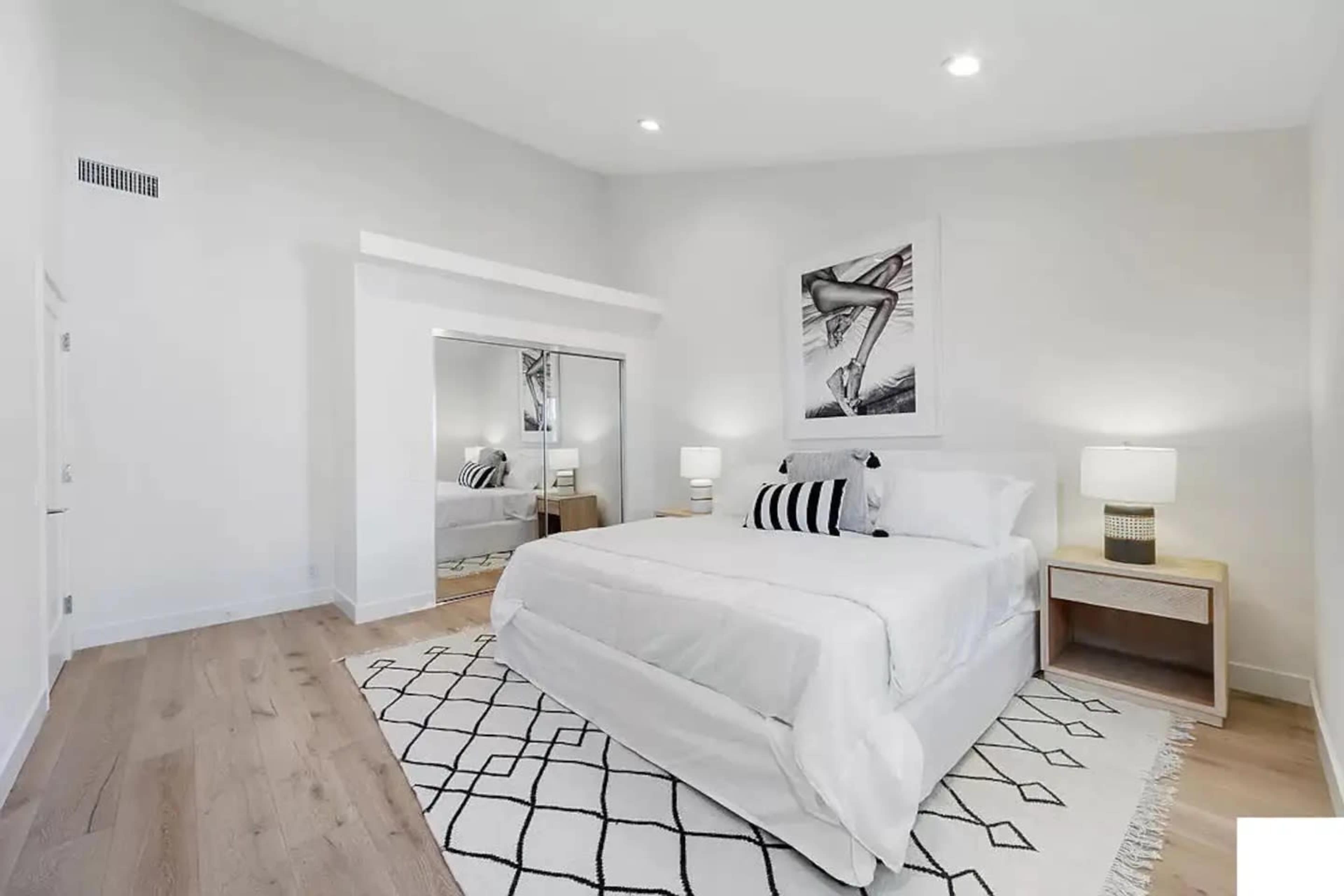 A brightly lit bedroom featuring a bed with white bedding, a large mirror, and framed artwork on the wall.