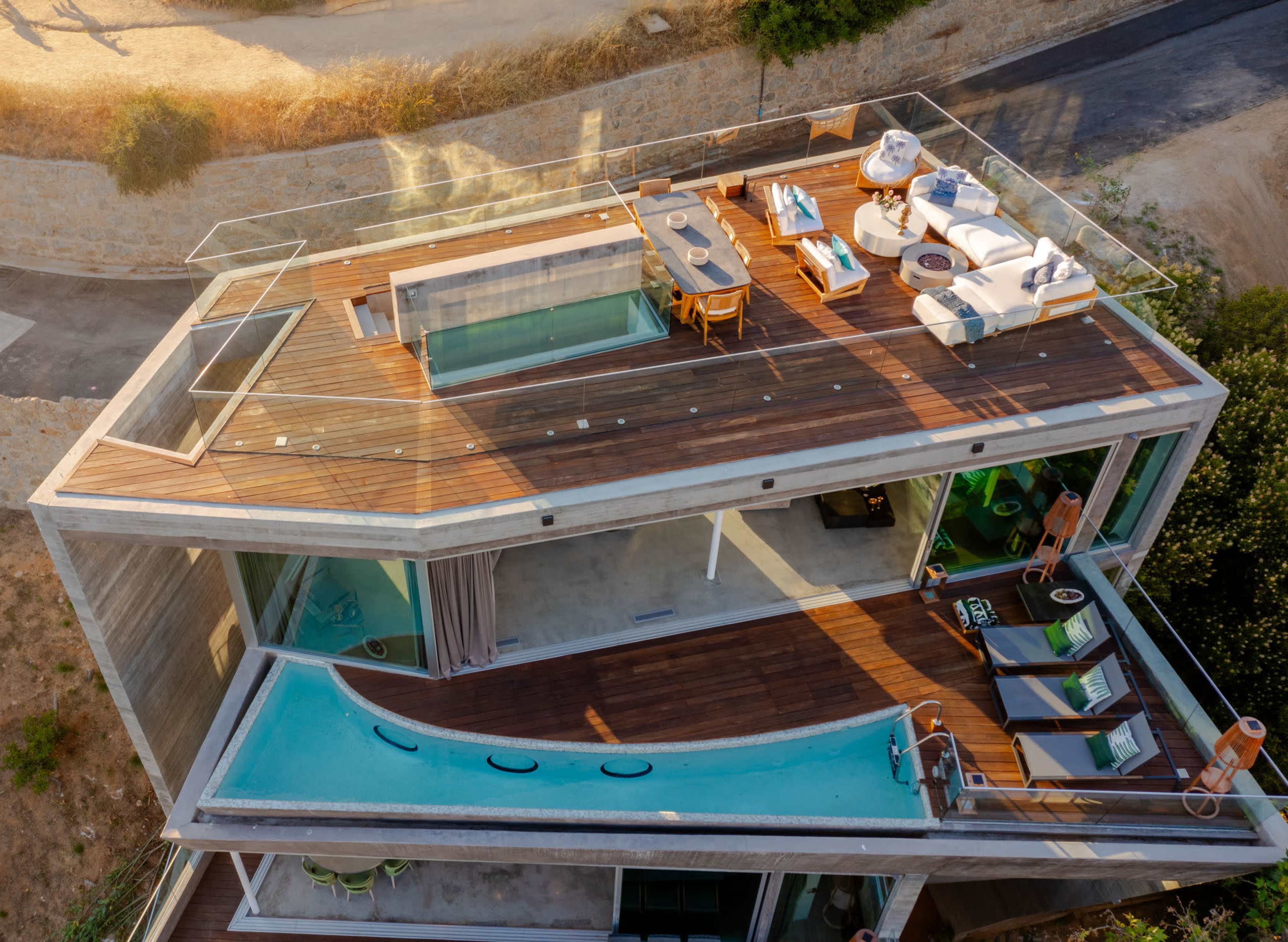 The image shows a modern house with a spacious outdoor deck featuring a dining area, lounge furniture, and two swimming pools on the roof.