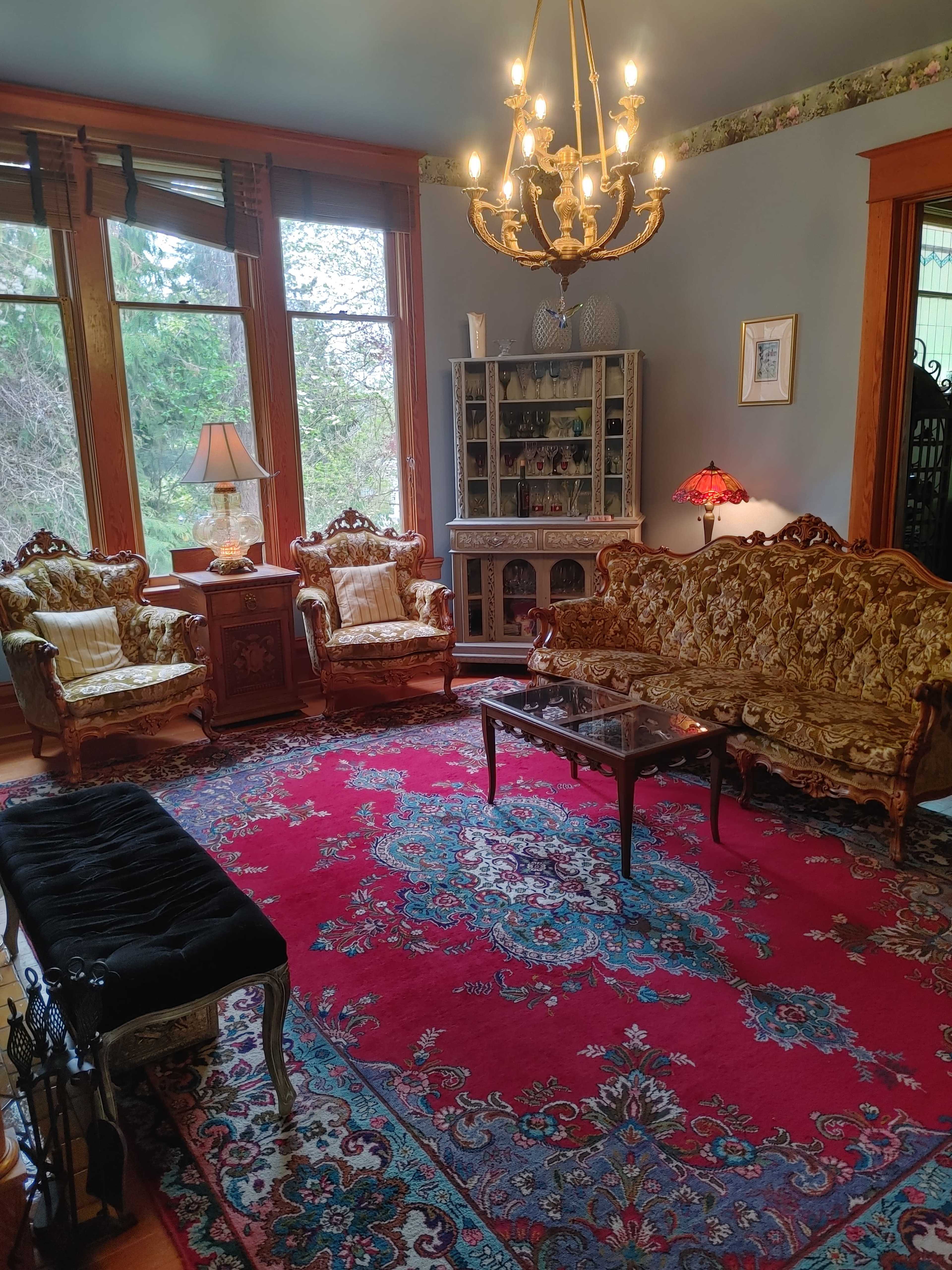 The room features ornate furniture, including two armchairs and a couch, surrounding a central coffee table on a large patterned rug, with a chandelier hanging from the ceiling.