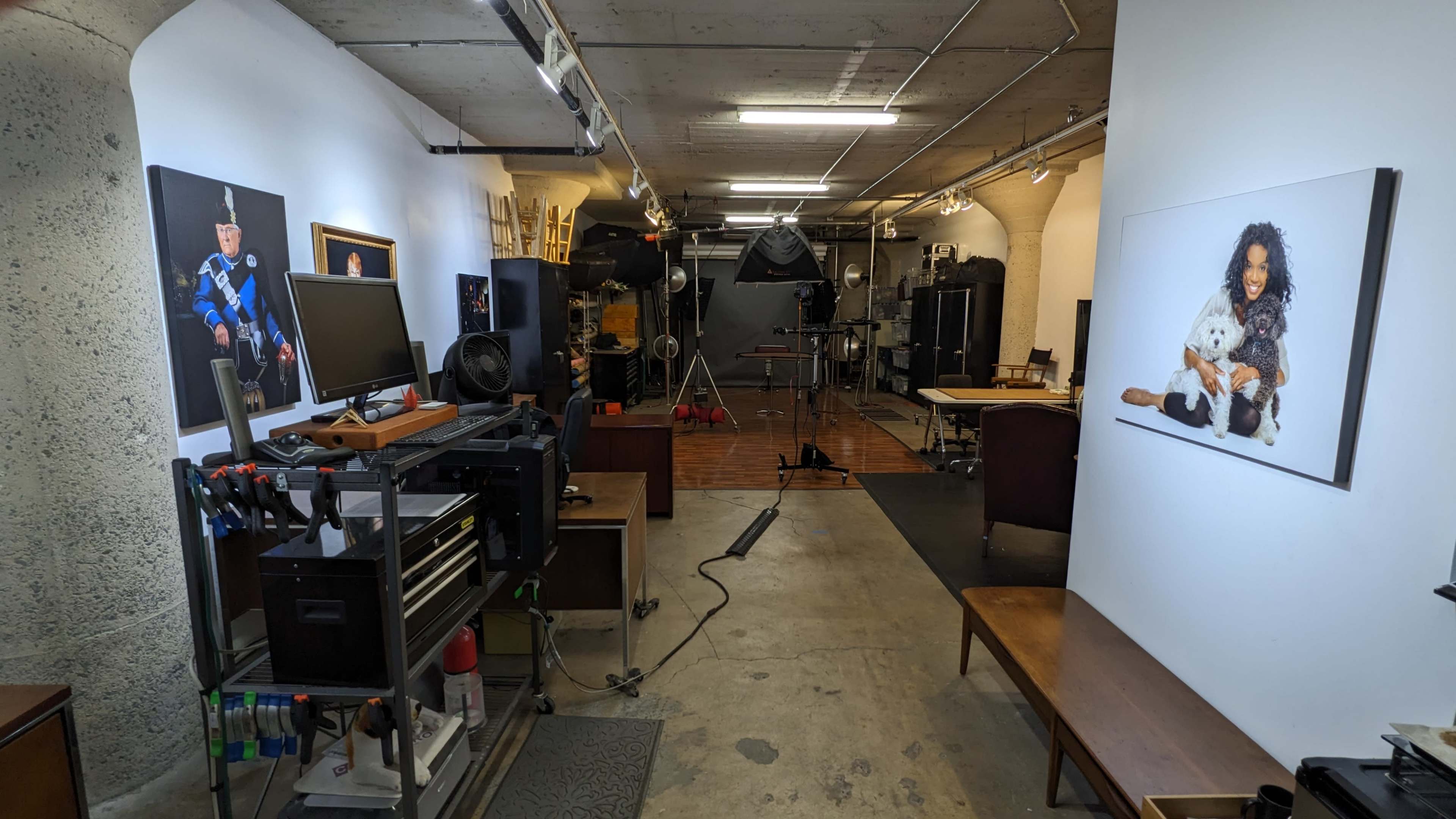 The image shows a photography studio with equipment, computers, and a small seating area, featuring two large framed photographs on the walls.