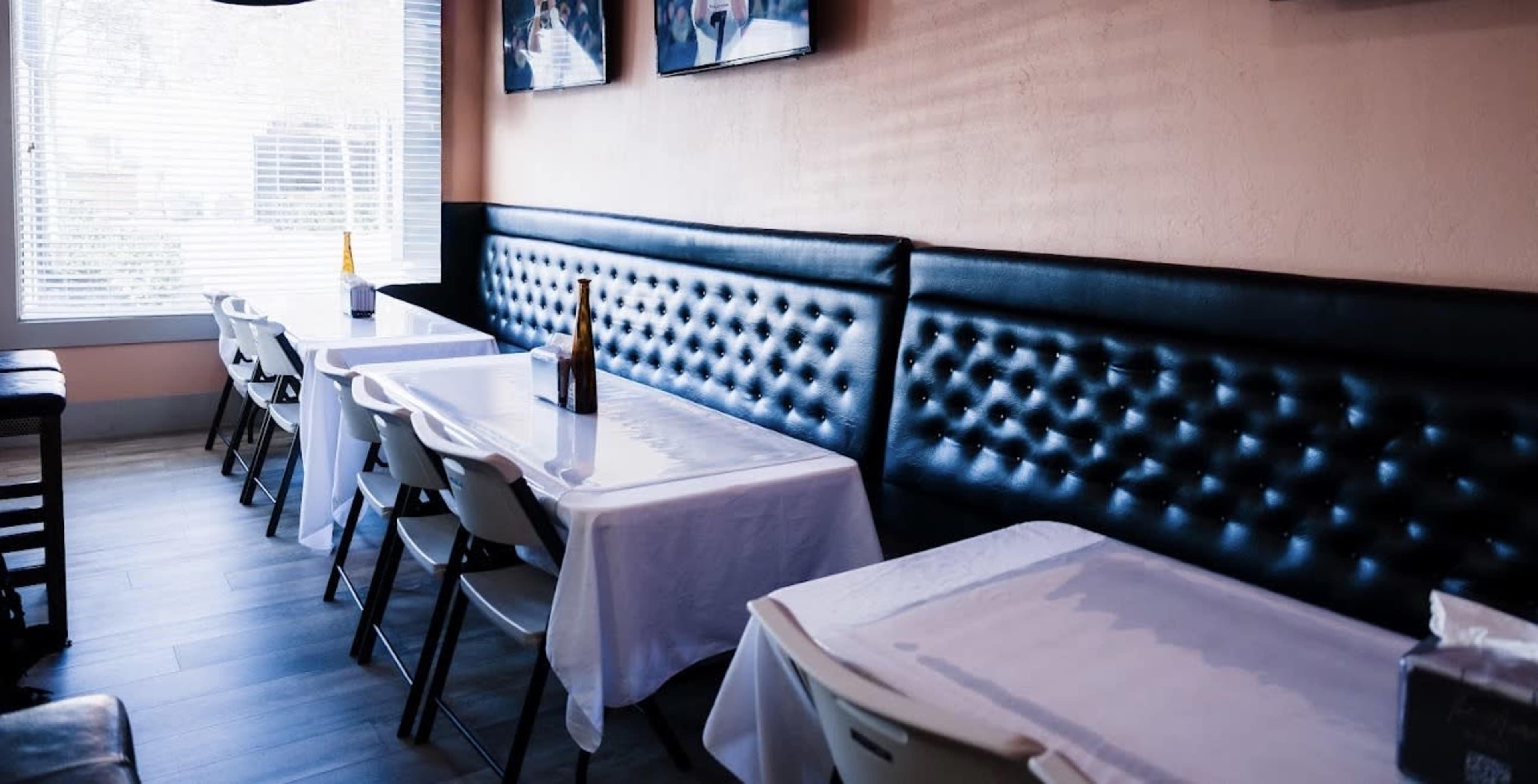A dining area features several white-covered tables along a black tufted bench under a pink wall with televisions.