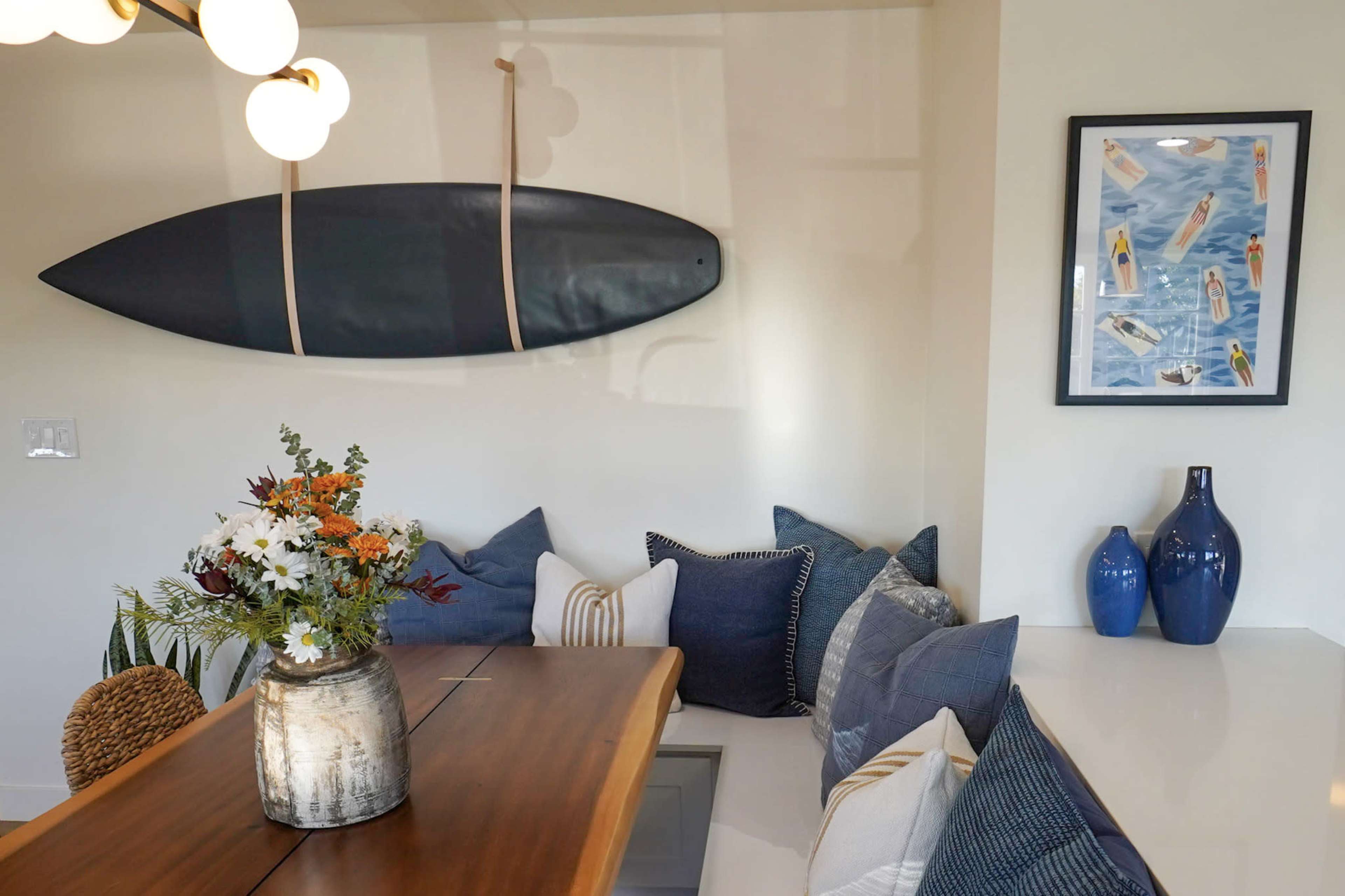 A wooden dining table is positioned near a wall featuring a surfboard, with decorative cushions and a floral arrangement on the table.