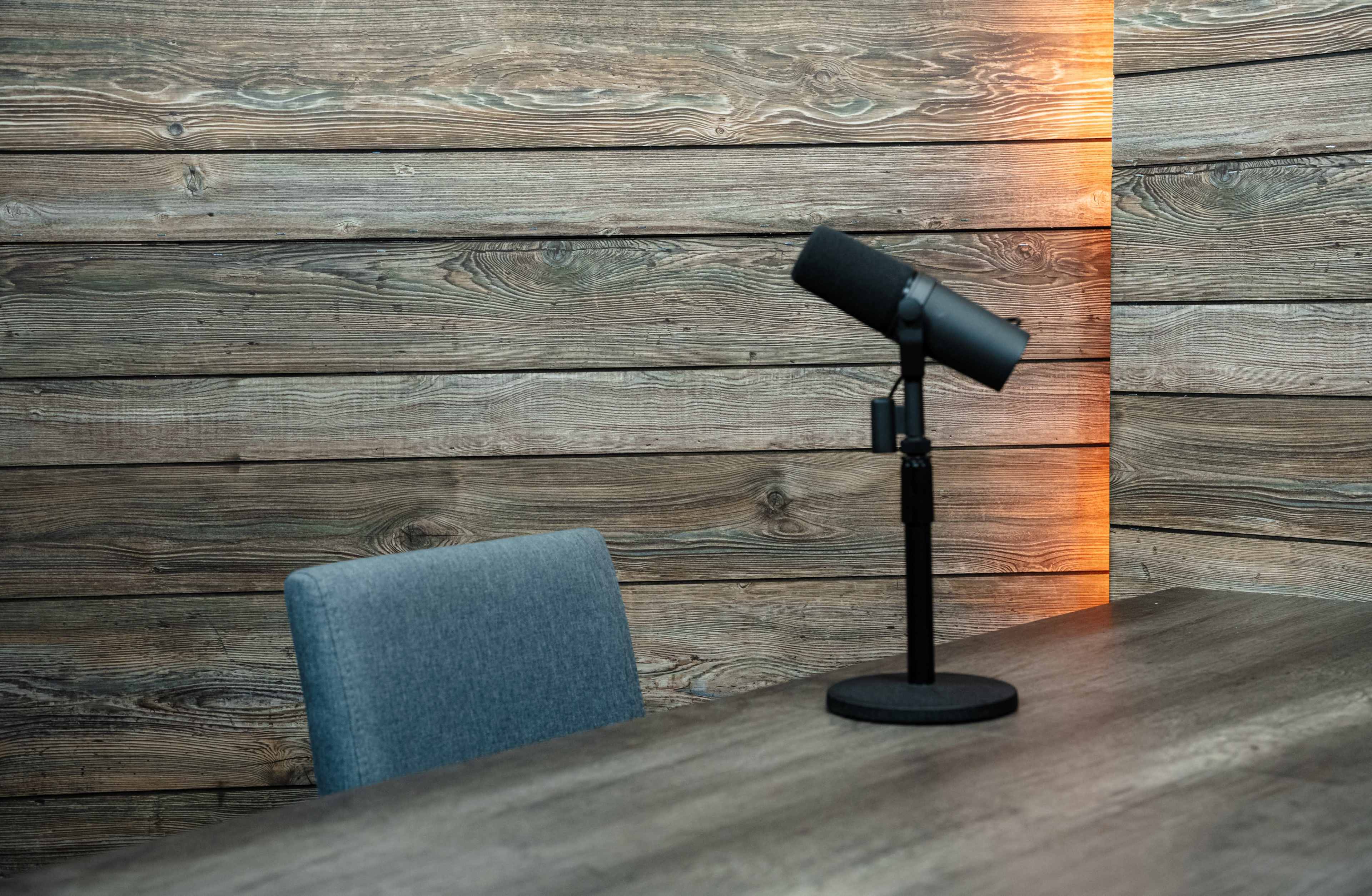 A microphone stands on a desk beside a blue chair against a backdrop of wooden paneling.