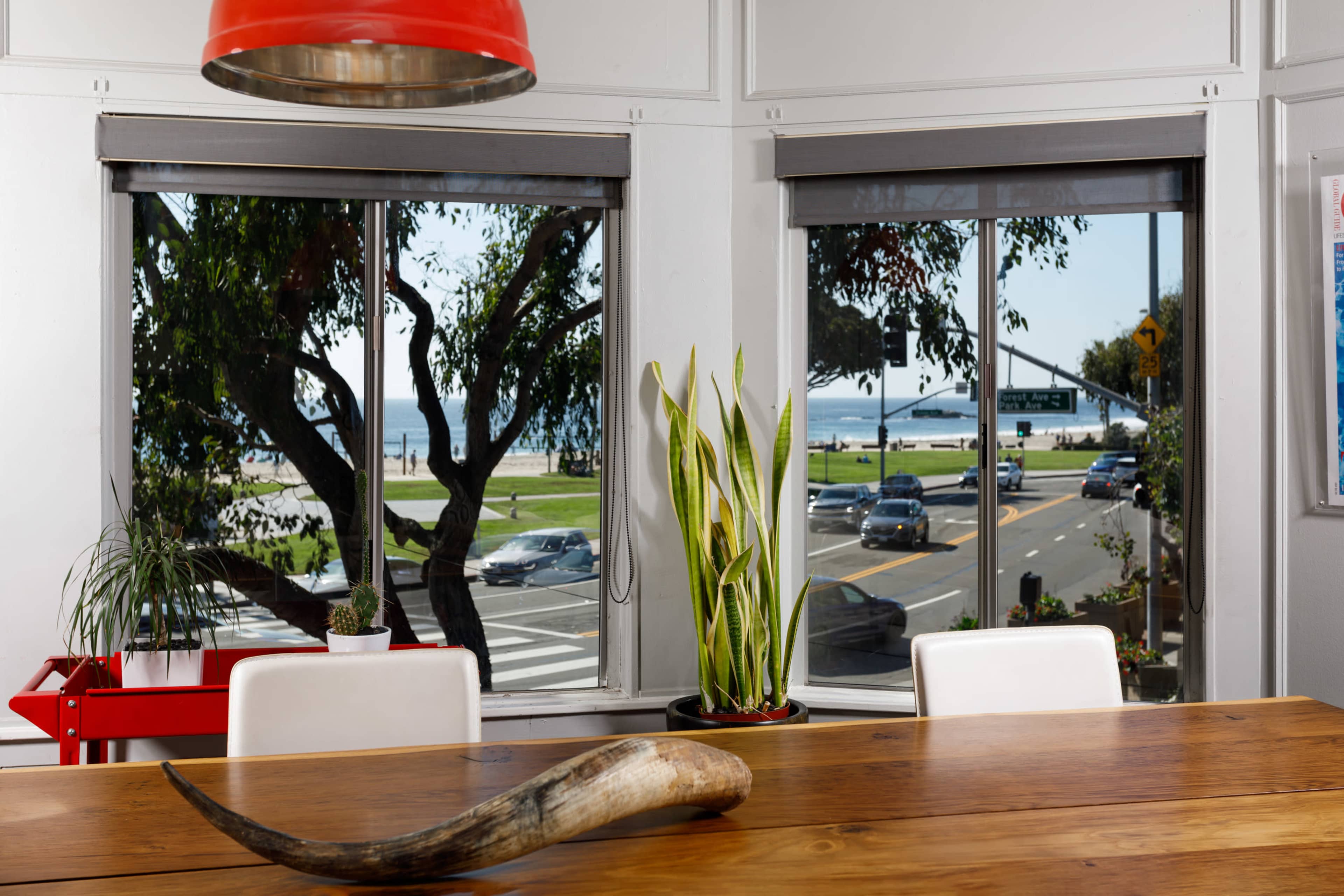 A dining area with a wooden table, a decorative horn, and large windows overlooking a street and waterfront scene with trees and vehicles.