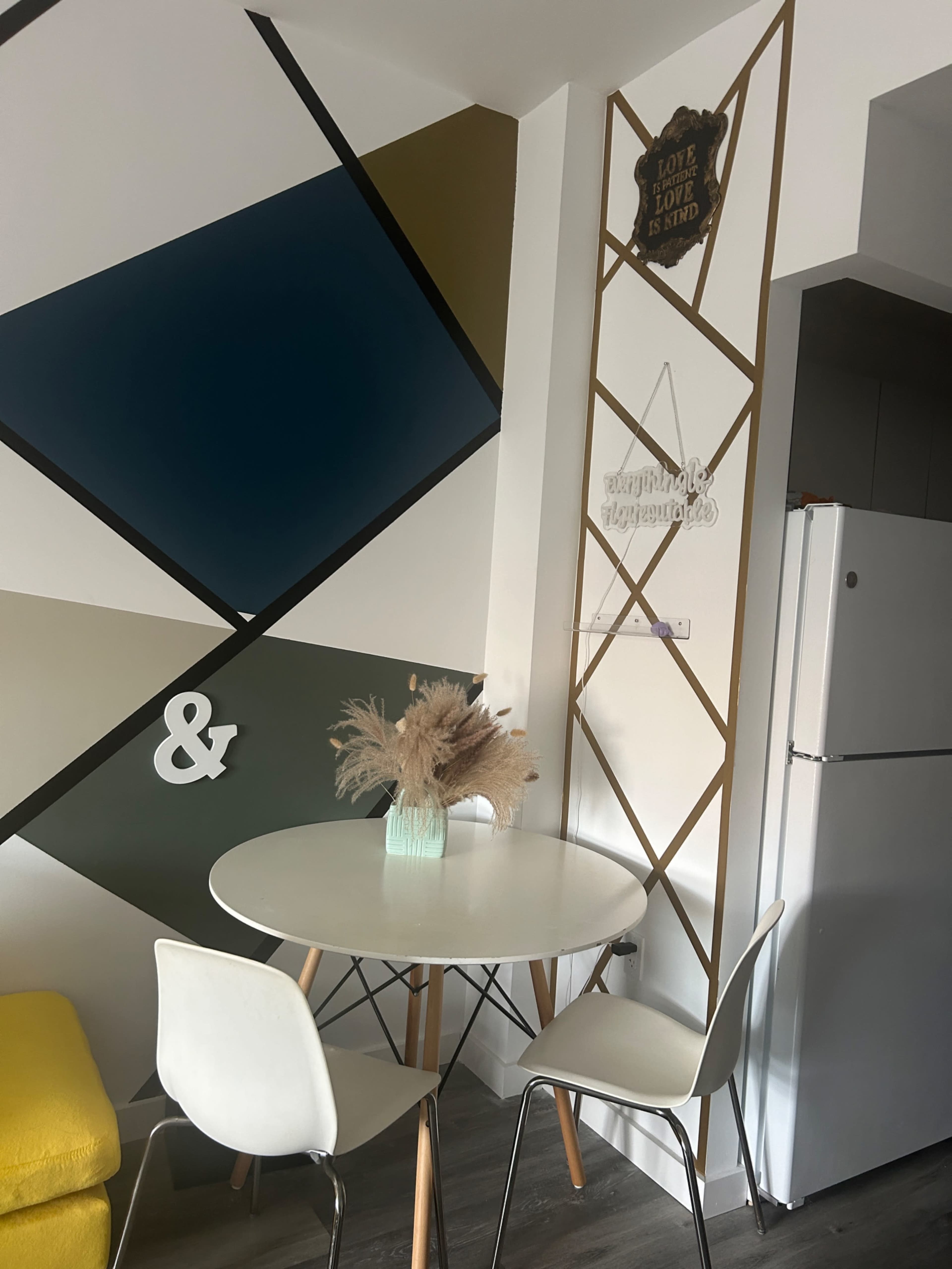 A small dining area features a round white table with chairs and a colorful geometric wall design in the background.