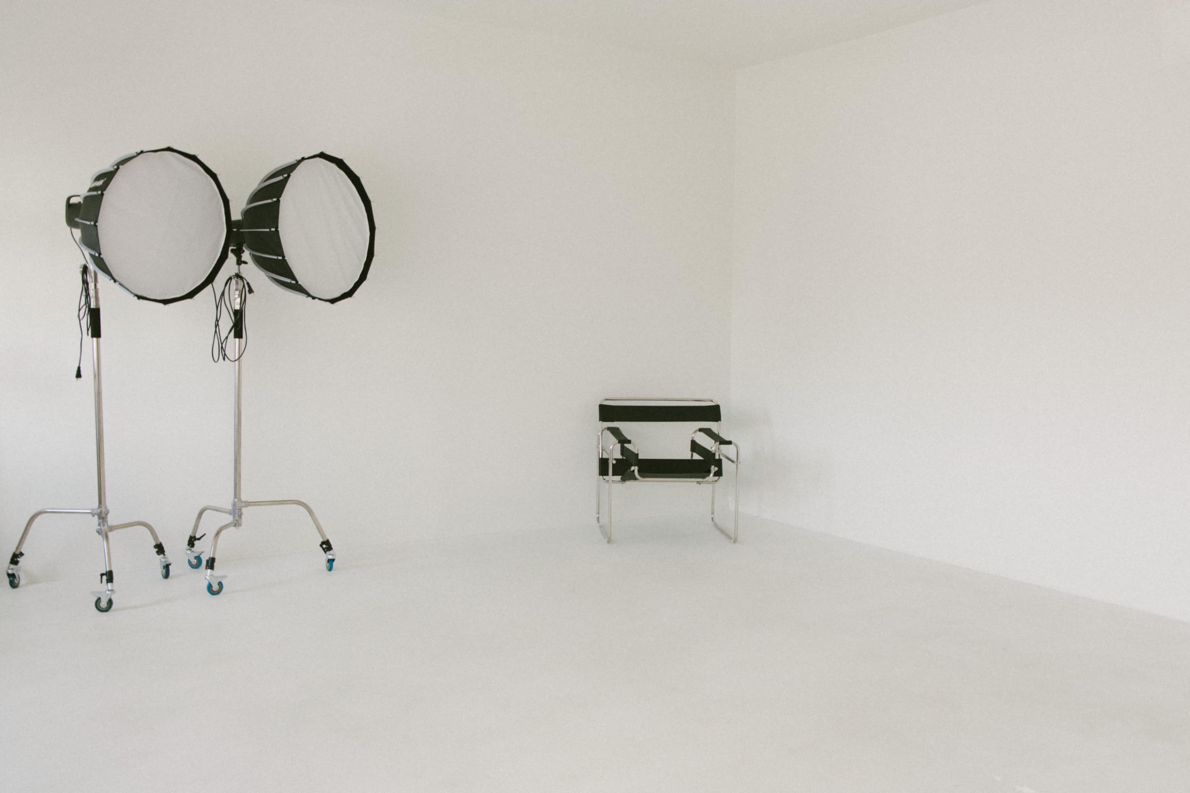 The image shows a minimalist, empty room with two studio lights on stands and a single chair against a plain white wall.