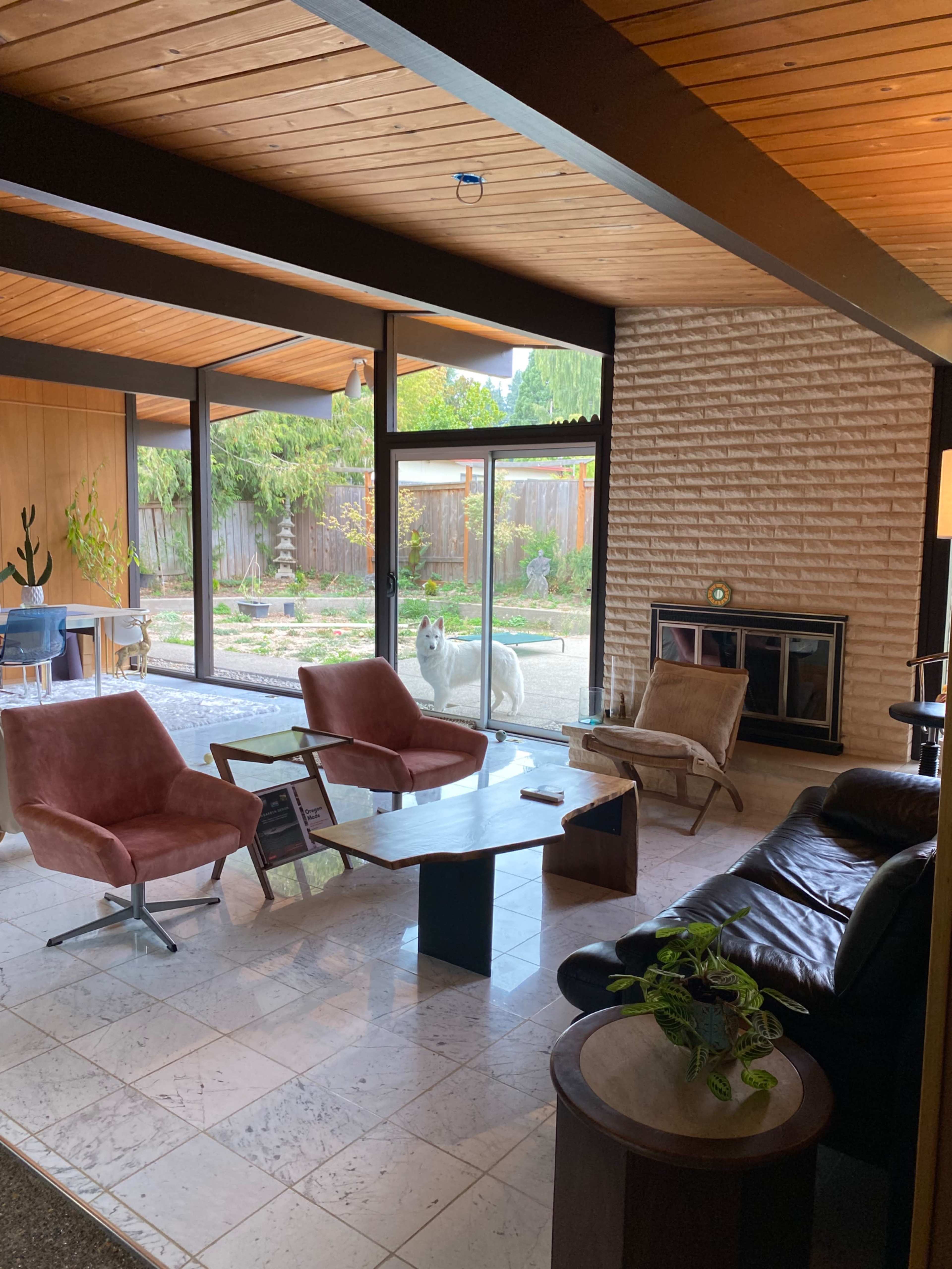 A mid-century modern living room with large windows, featuring a mix of seating options, a coffee table, and a view of a garden outside.