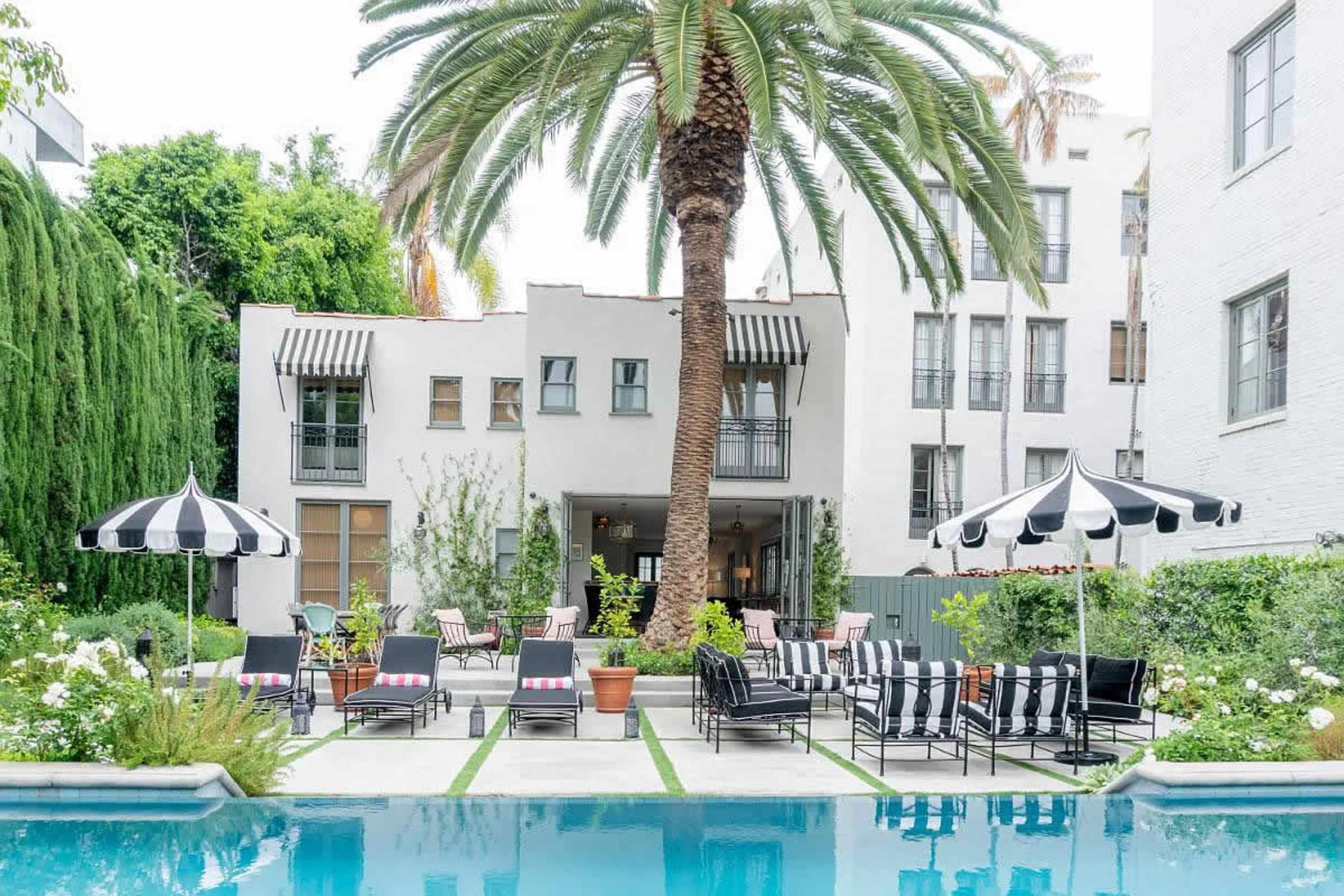 The image shows a pool area surrounded by lounge chairs and striped umbrellas, with a backdrop of a white building and palm trees.
