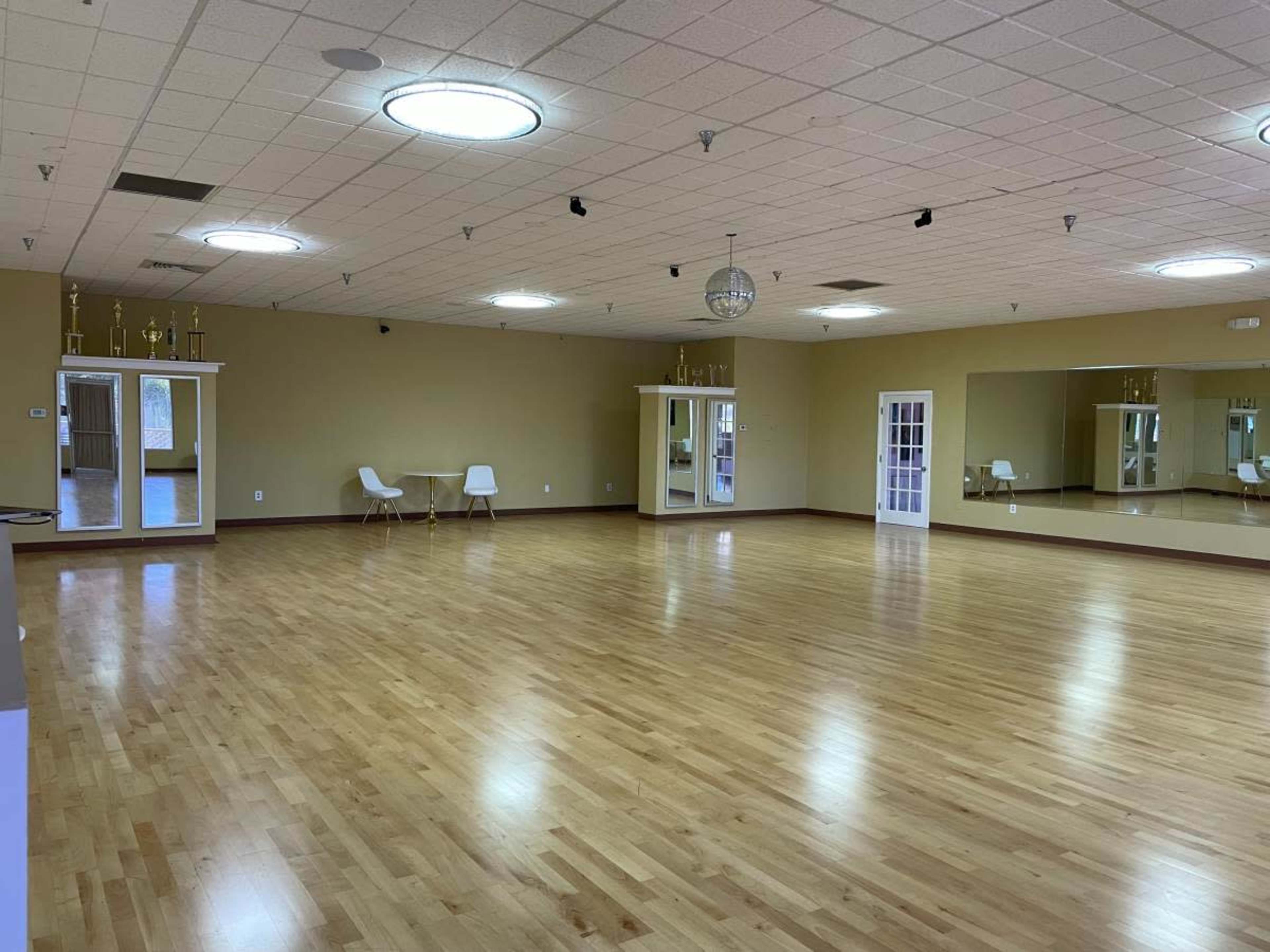 A large, empty dance studio with wooden floors, mirrors, and a few chairs set against the walls.