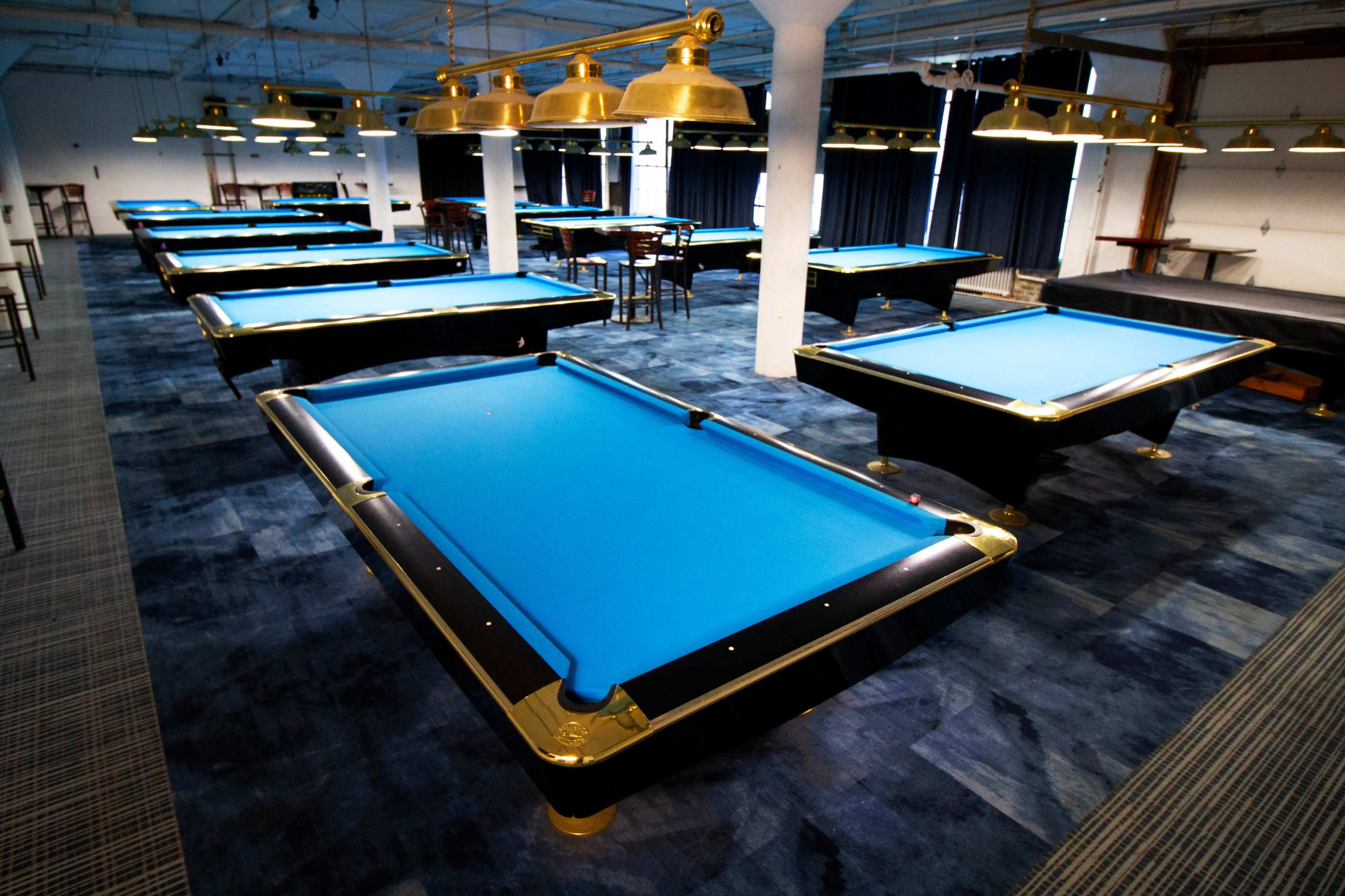 A spacious billiards room with multiple pool tables featuring blue felt tops and industrial-style lighting.