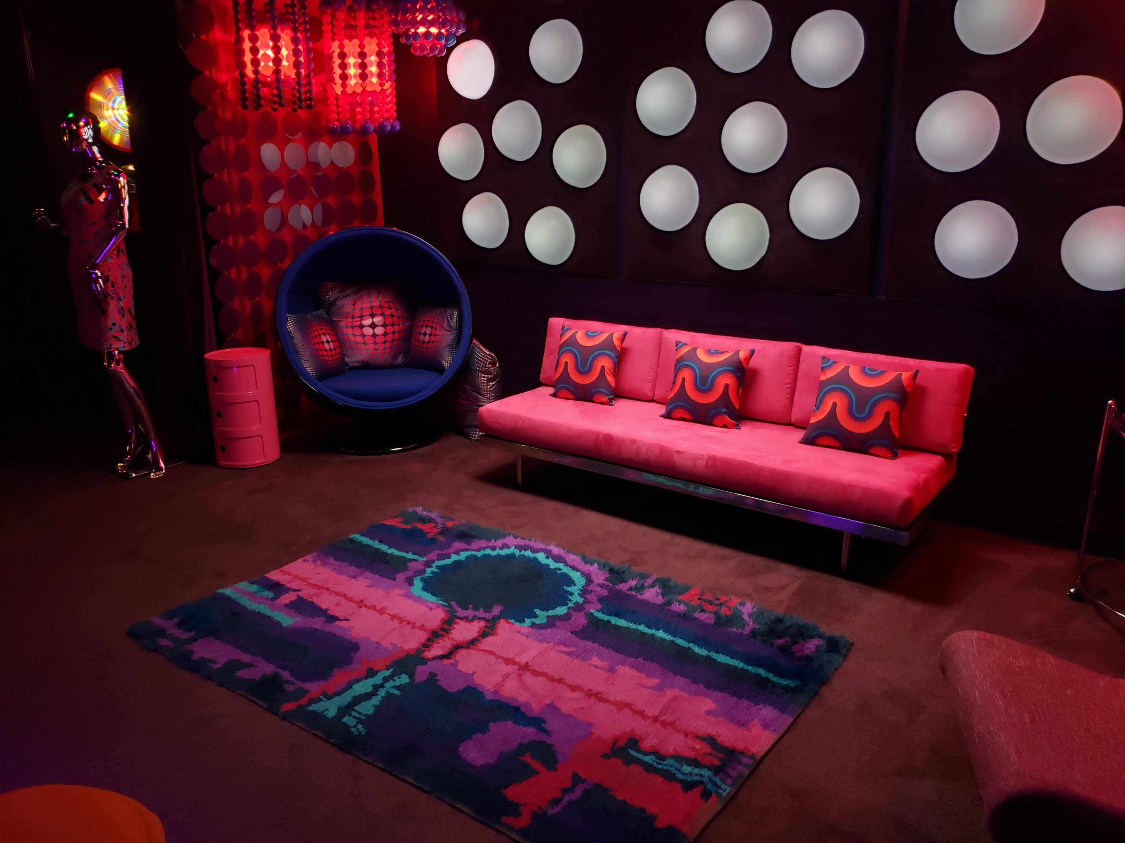 The room features a pink sofa with patterned cushions, a blue chair, colorful lighting, and a large rug with a tree design against a backdrop of circular wall decorations.