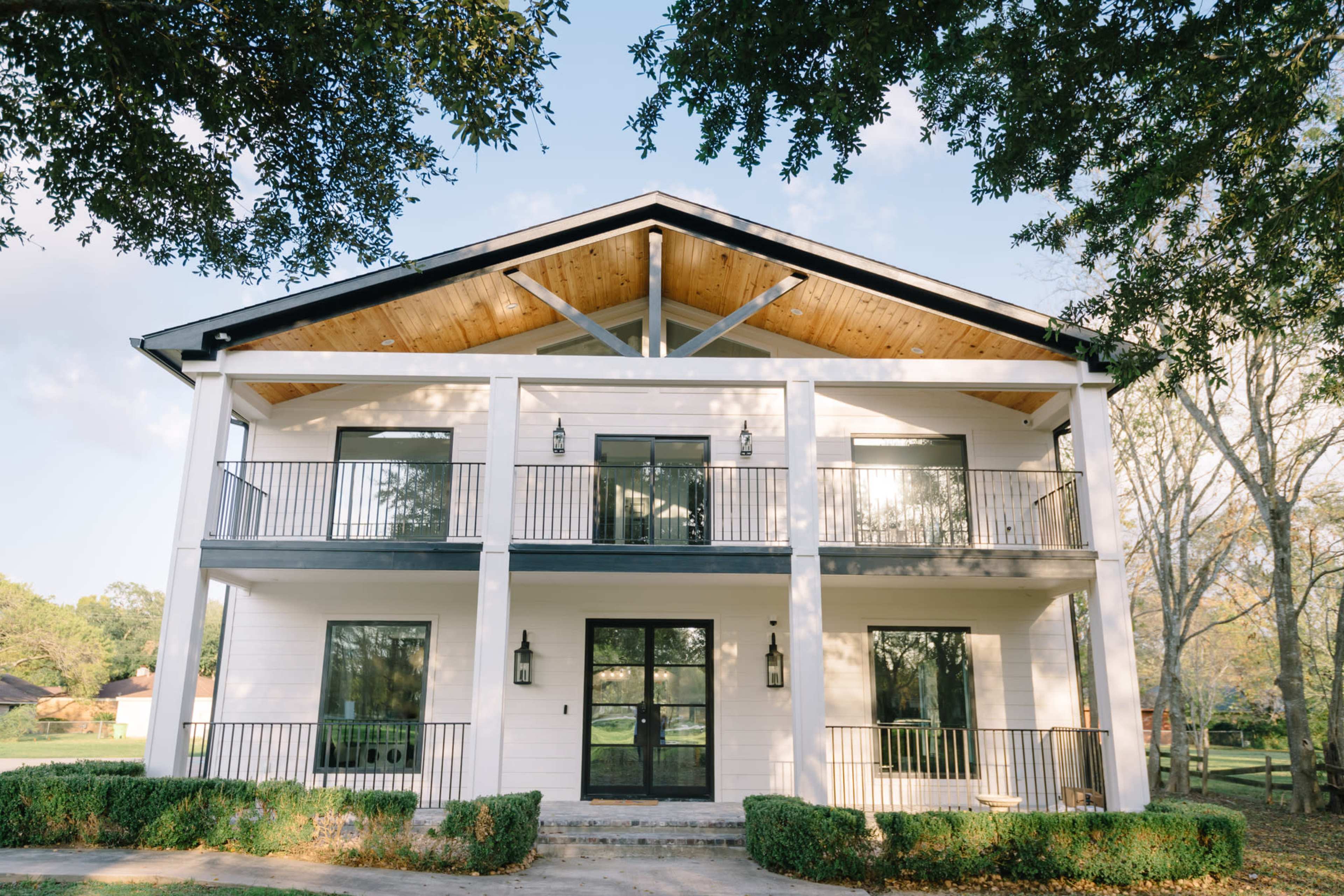 A two-story modern house features a white exterior, large black-framed windows, and a peaked roof with wooden beams.