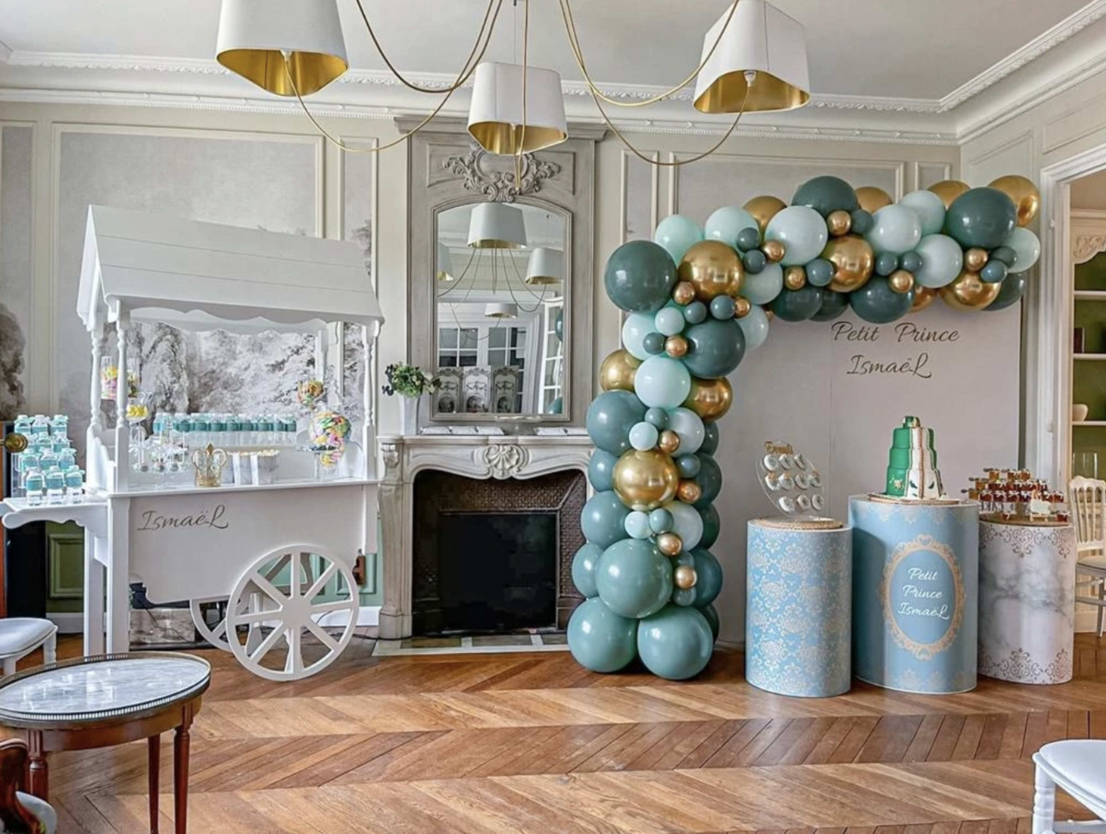 A decorated indoor space featuring a balloon arch in shades of teal and gold, a small beverage cart, and a dessert table.