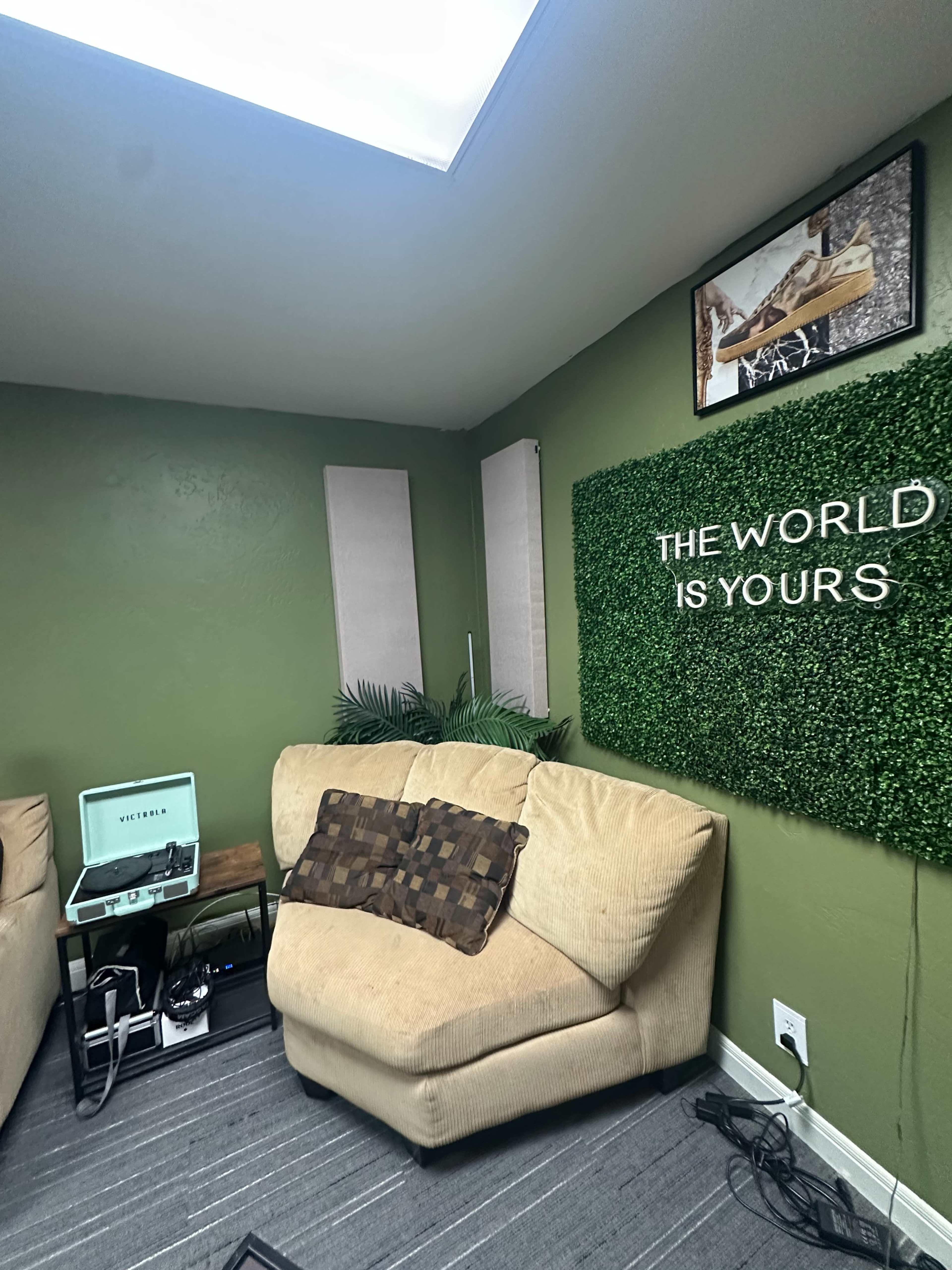 The image shows a cozy corner of a room with a green wall adorned with the phrase "THE WORLD IS YOURS," a beige couch, and a record player next to it.