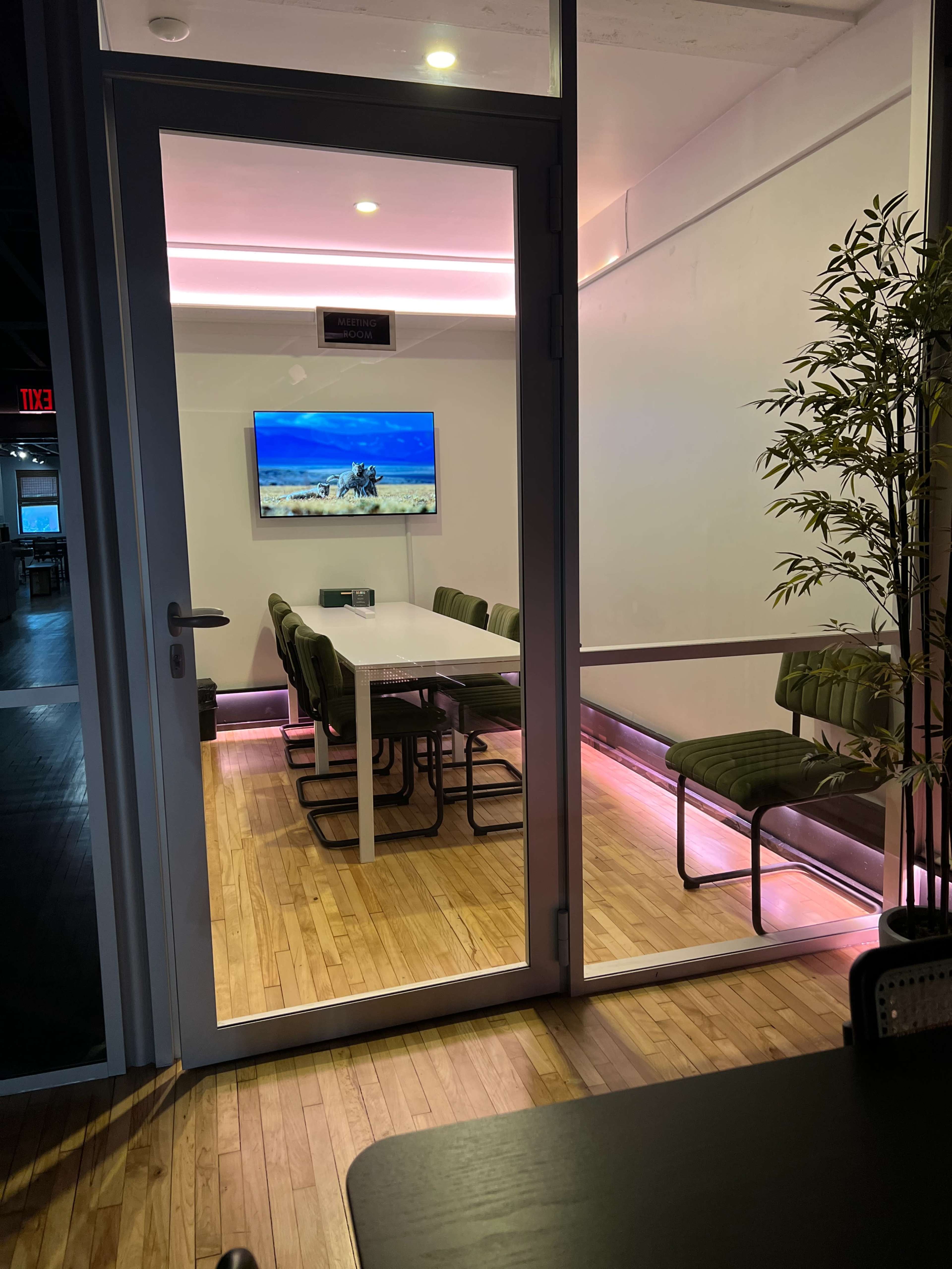 A modern conference room features a large table surrounded by green chairs and a wall-mounted TV, illuminated by pink LED lights.