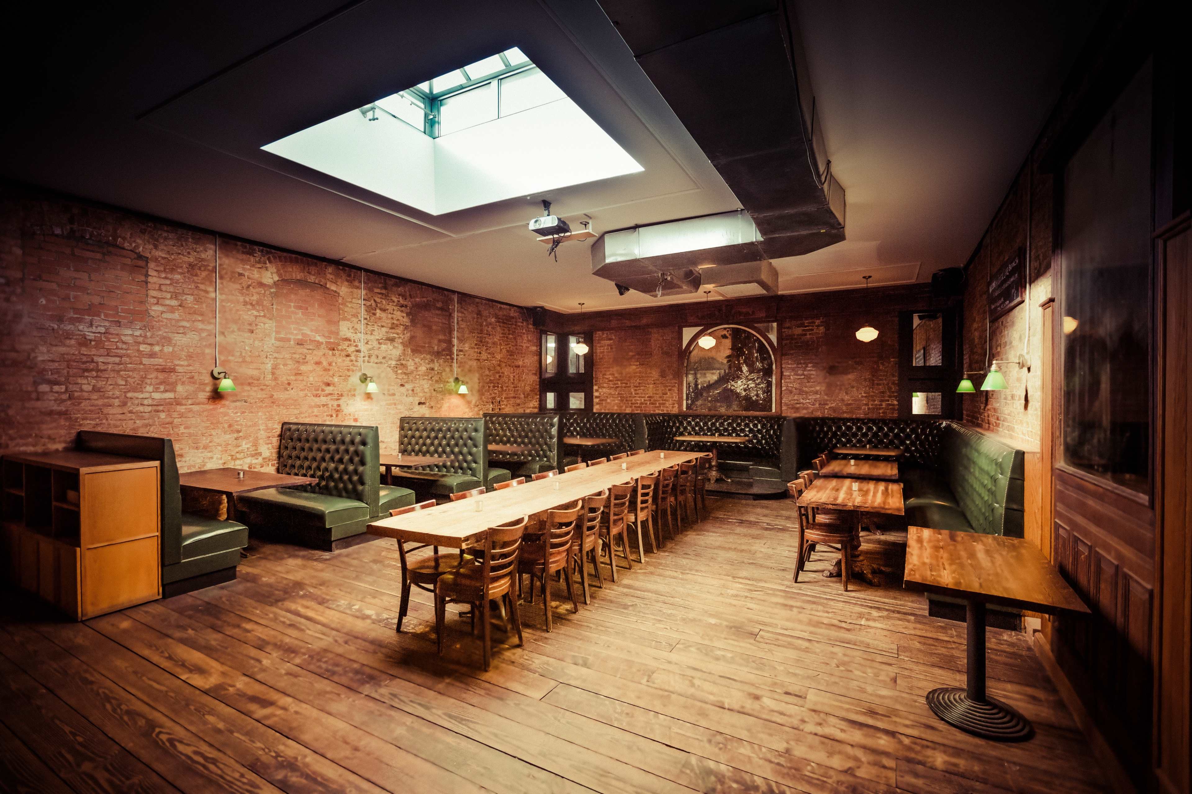 The interior of a restaurant features exposed brick walls, wooden floors, and a series of long wooden tables with green upholstered seating along the walls, illuminated by green pendant lights.