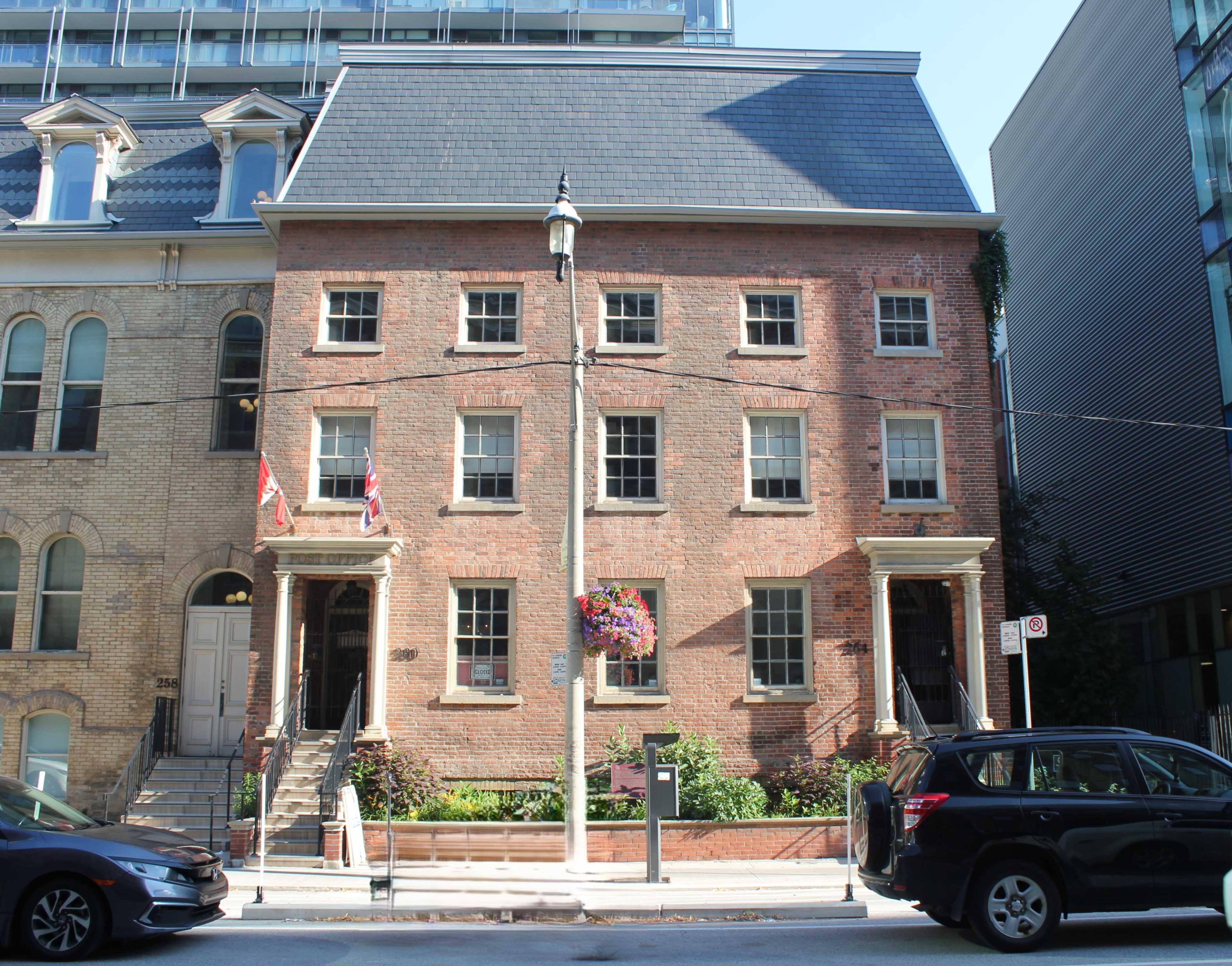 The image shows a historic brick building with a symmetrical facade, featuring four windows on the ground floor, two British flags, and a garden area along the front.