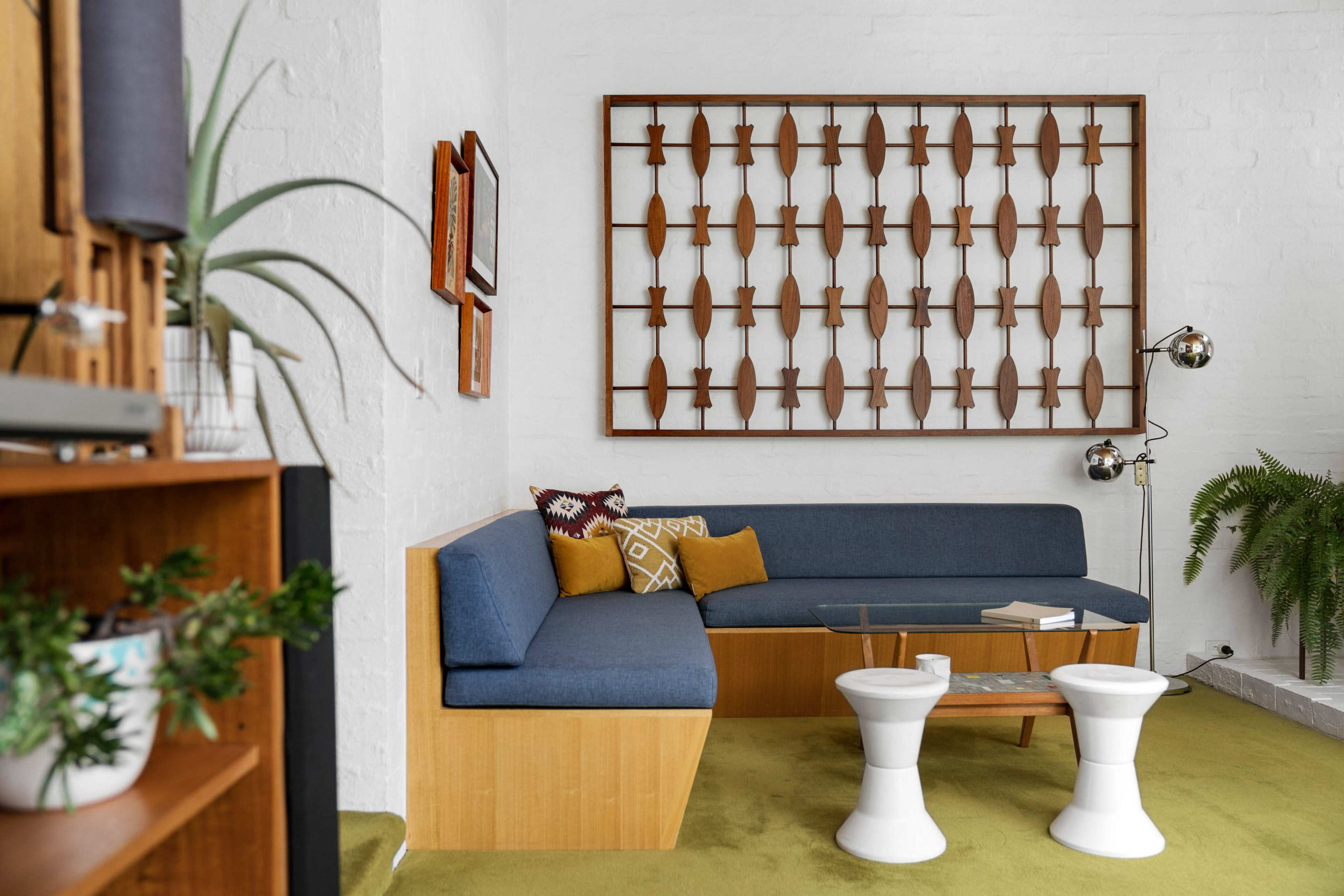 The image shows a modern living room featuring a blue sectional sofa, a glass coffee table, and a decorative wall art piece made of wooden elements, surrounded by plants and bookshelves.
