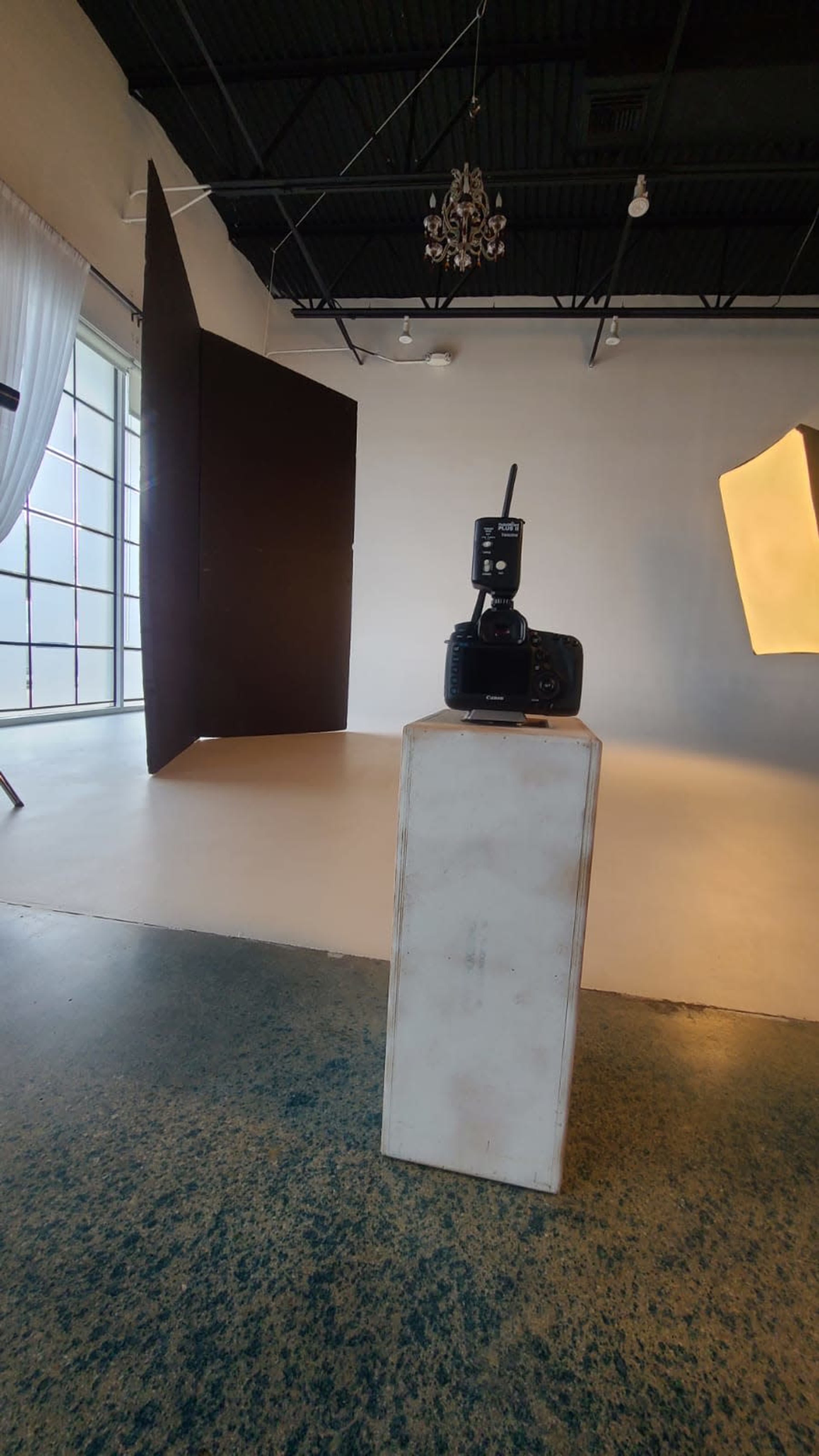 The image shows a photography studio with a camera on a pedestal in the foreground and a large light source and backdrop in the background.