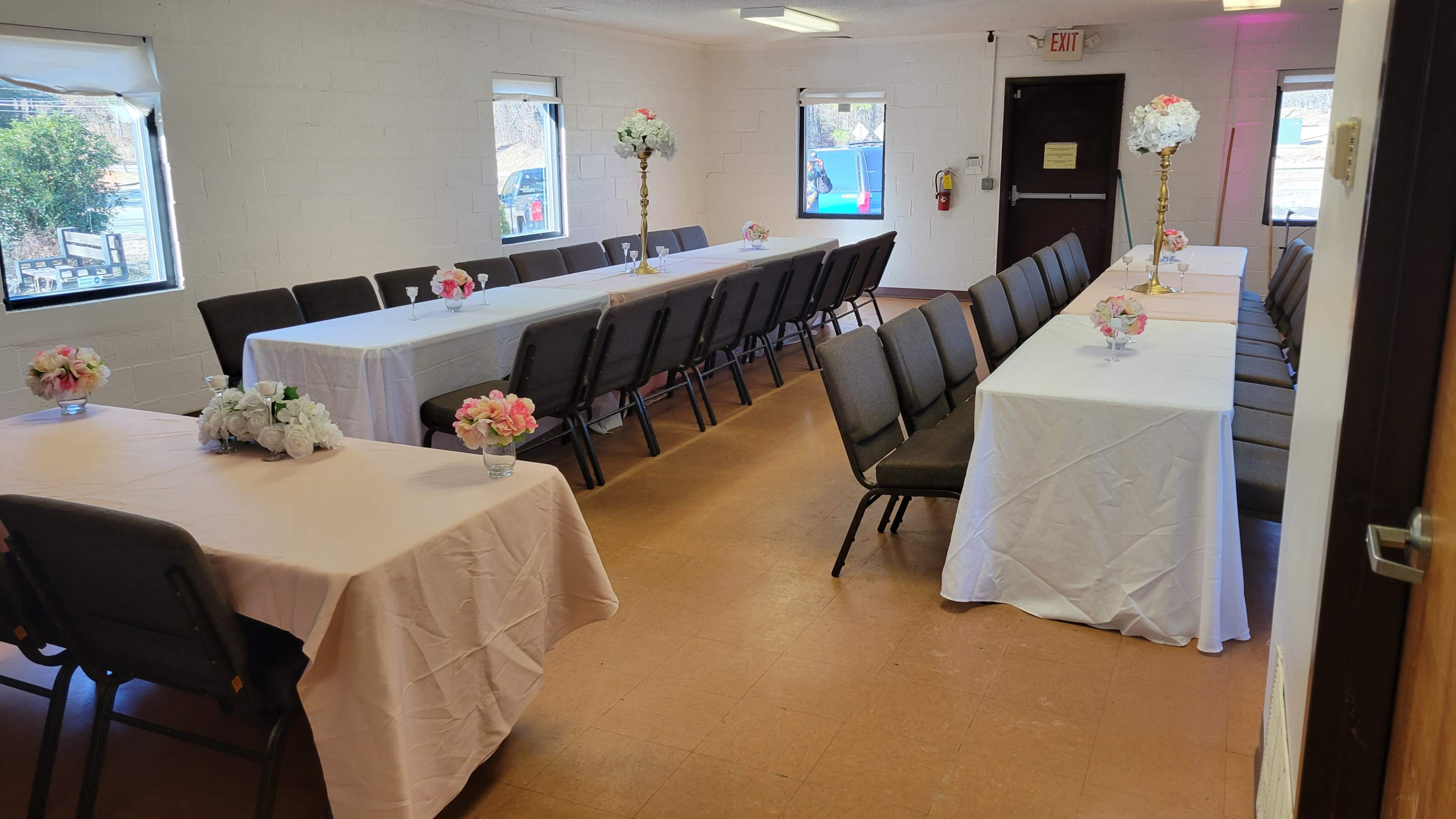 The image shows a room set up for an event with two long tables covered in white and pink tablecloths, each adorned with floral centerpieces, and surrounded by gray chairs.