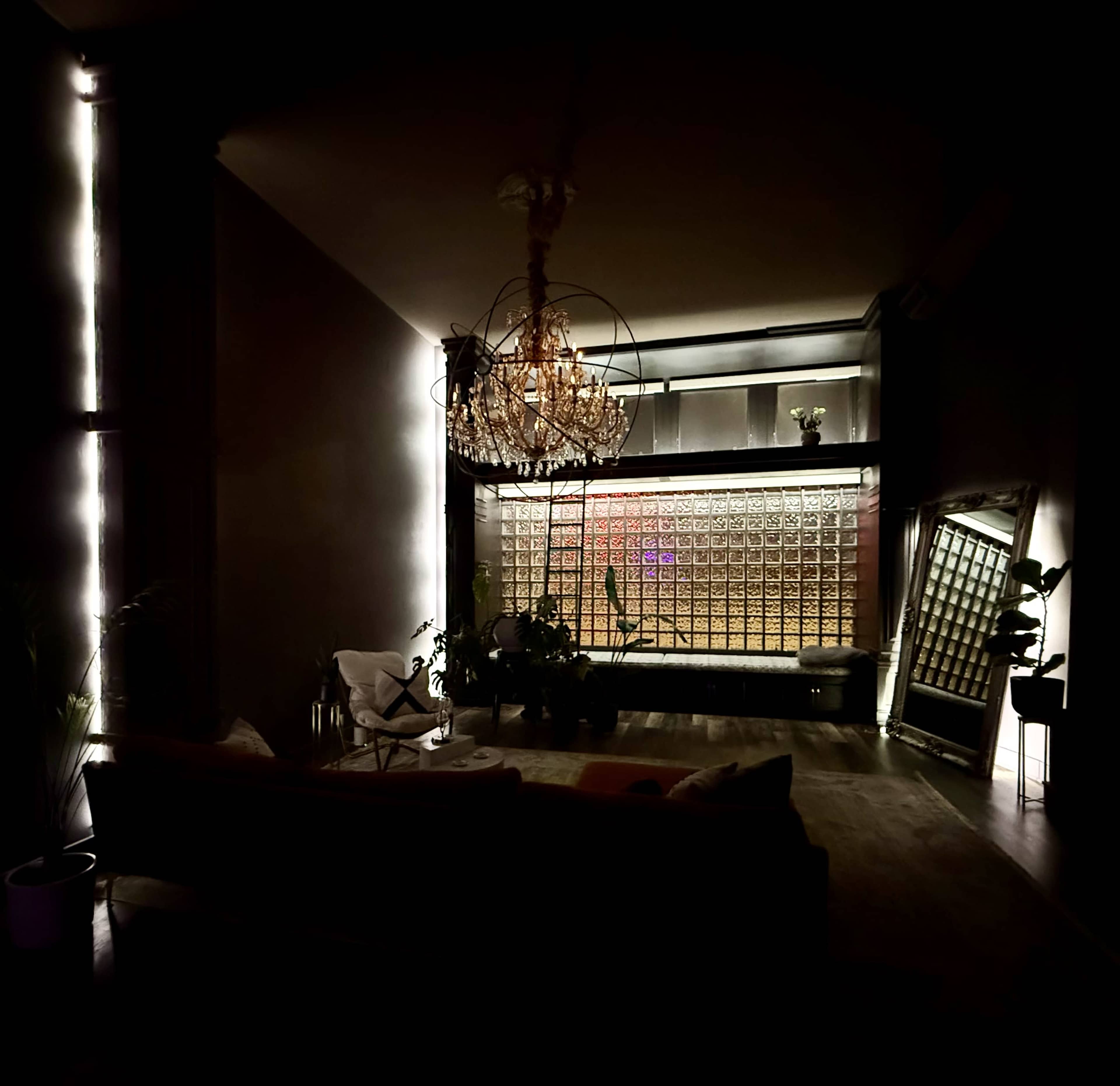 The image shows a dimly lit room featuring a chandelier, a sofa, a mirror, and a wall adorned with decorative glass blocks.