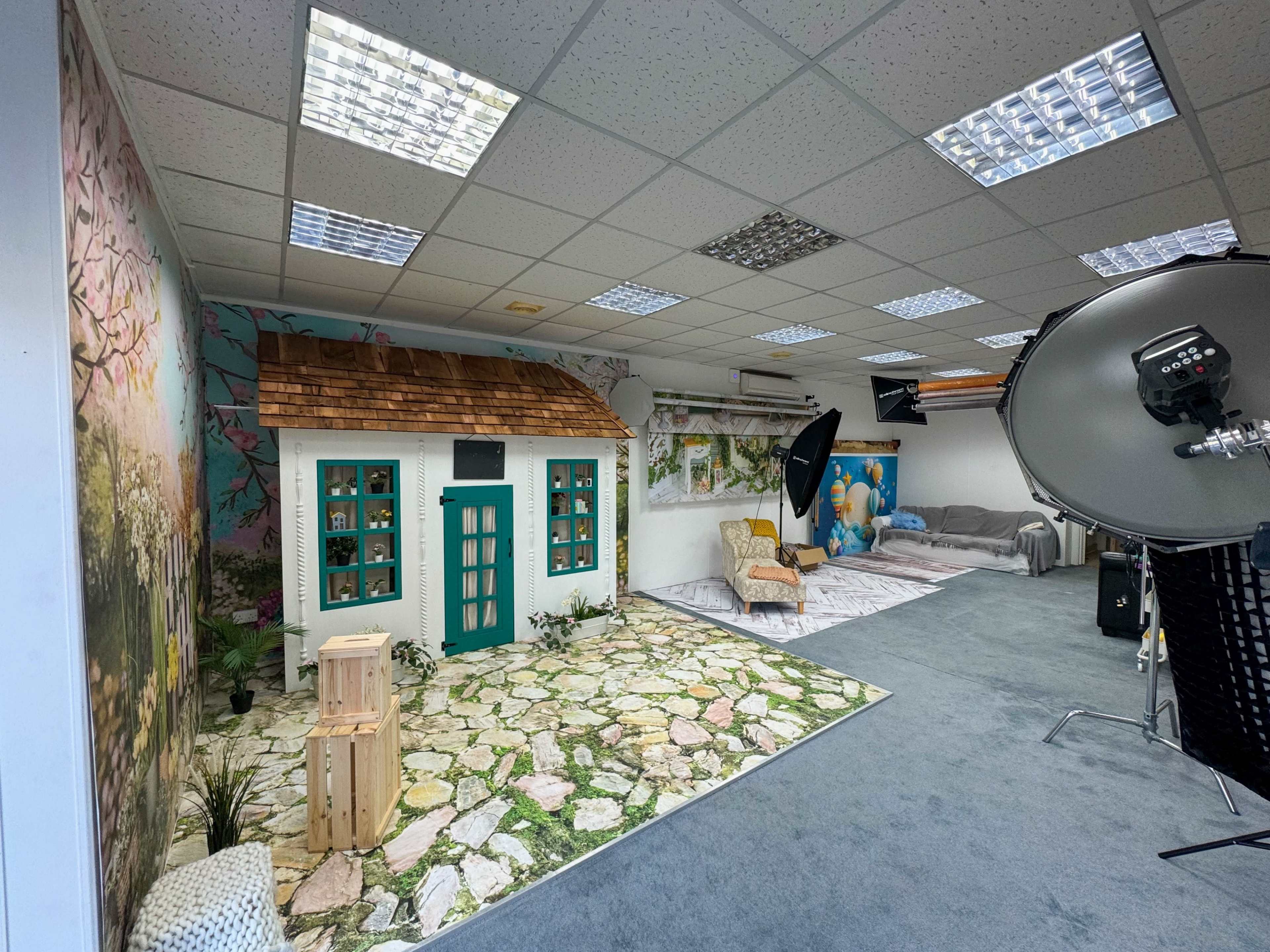 A spacious studio with a decorative backdrop featuring a small white cottage and a stone pathway, alongside a seating area with a couch and photography equipment.