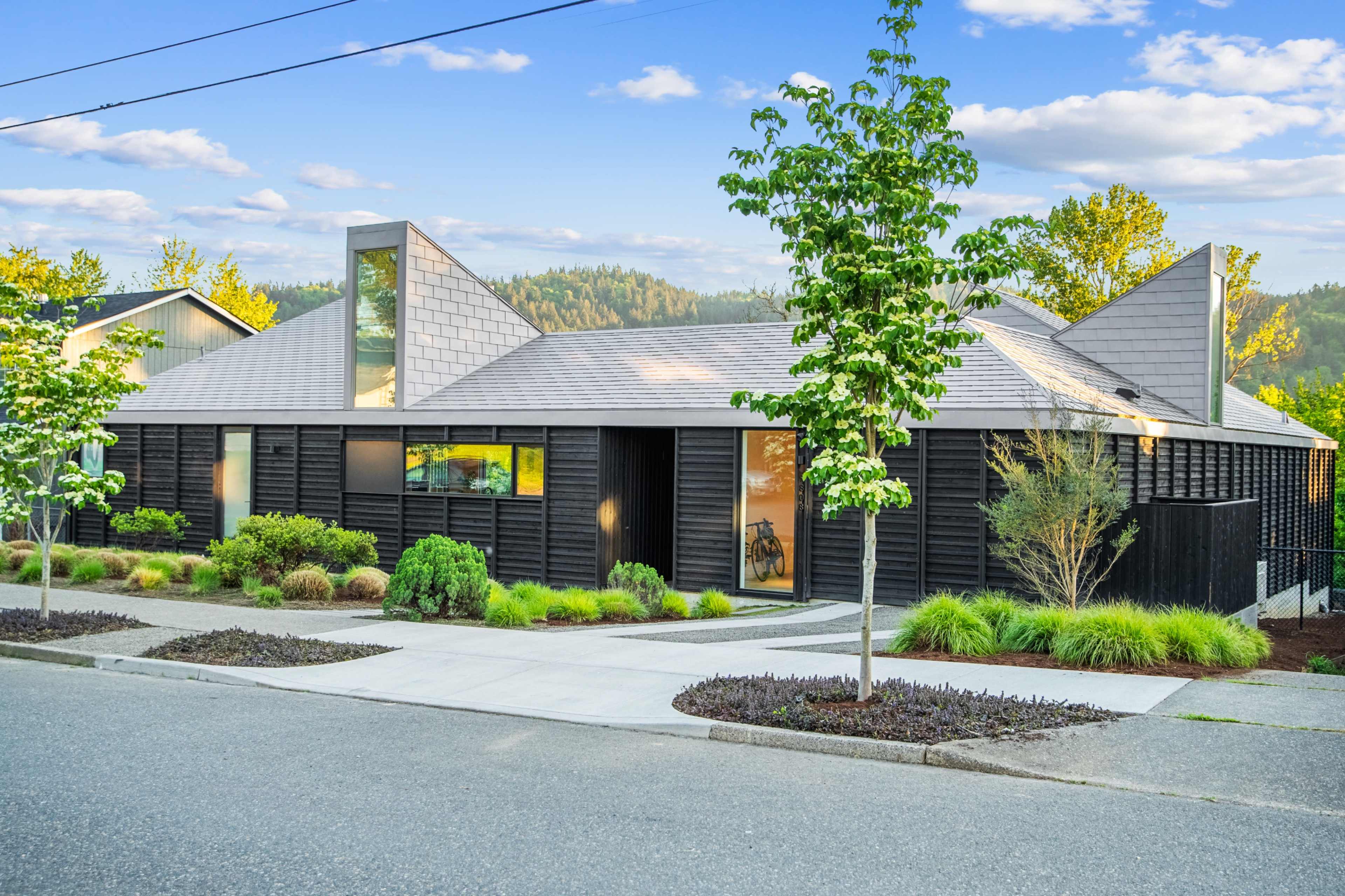 A modern black-clad house with angular roofs and landscaped greenery is situated along a paved street.