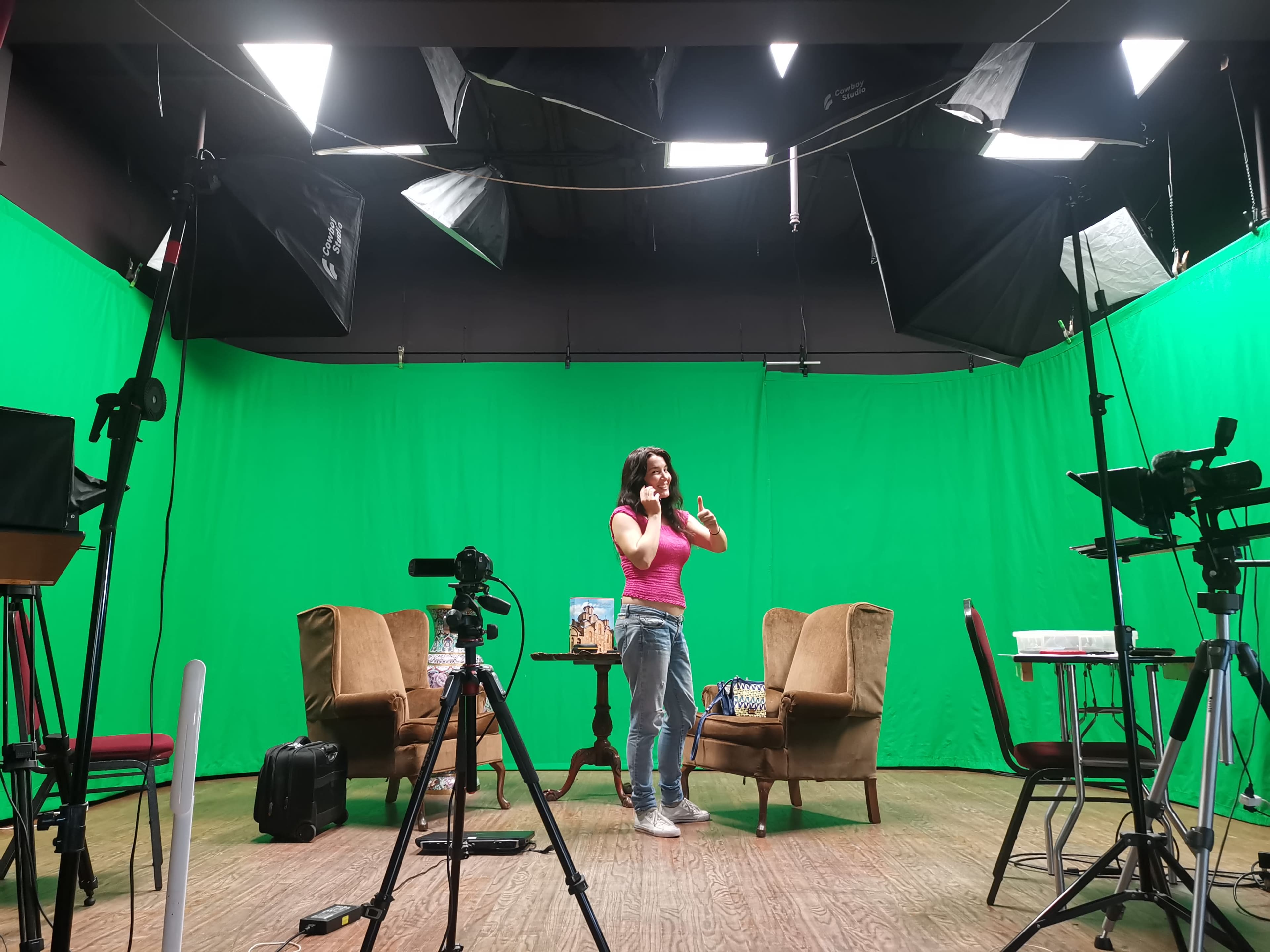 A woman stands in front of a green screen set, surrounded by cameras and lights, with two chairs positioned nearby.