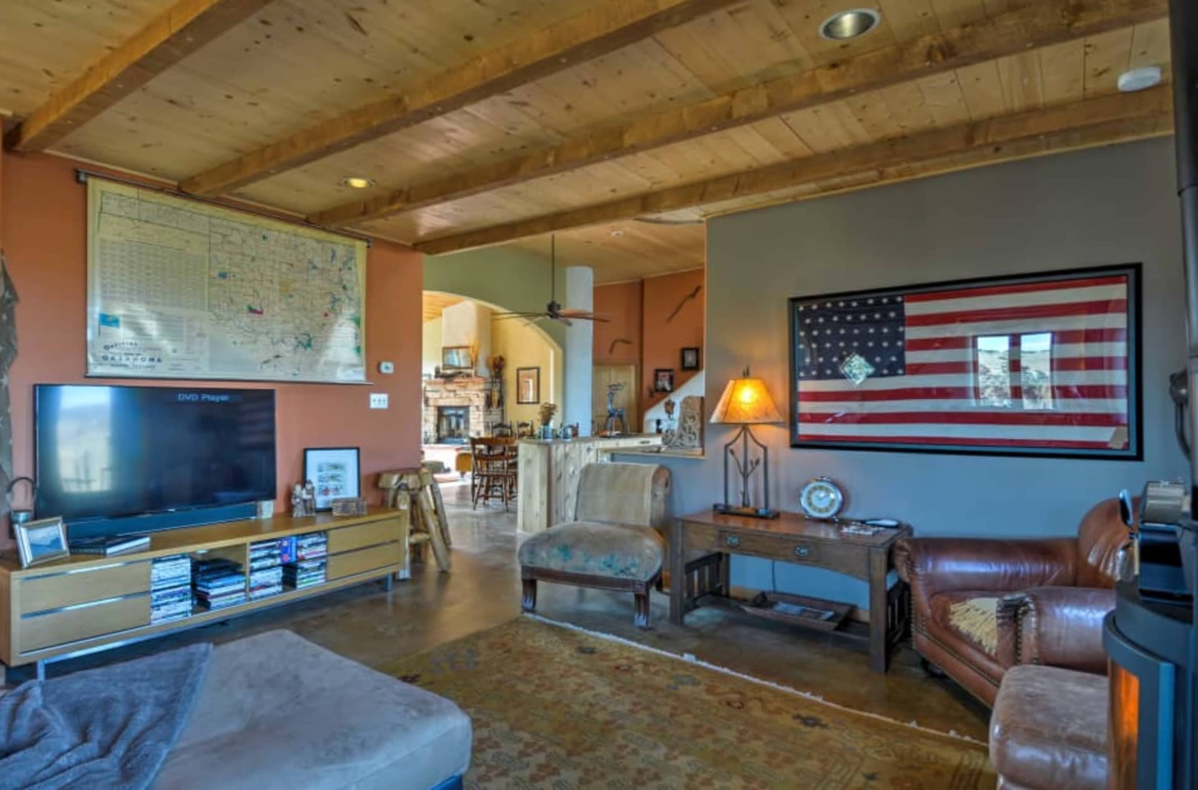 The living room features a wooden beam ceiling, a large television, a vintage map on the wall, and an American flag framed beside a comfortable seating area.