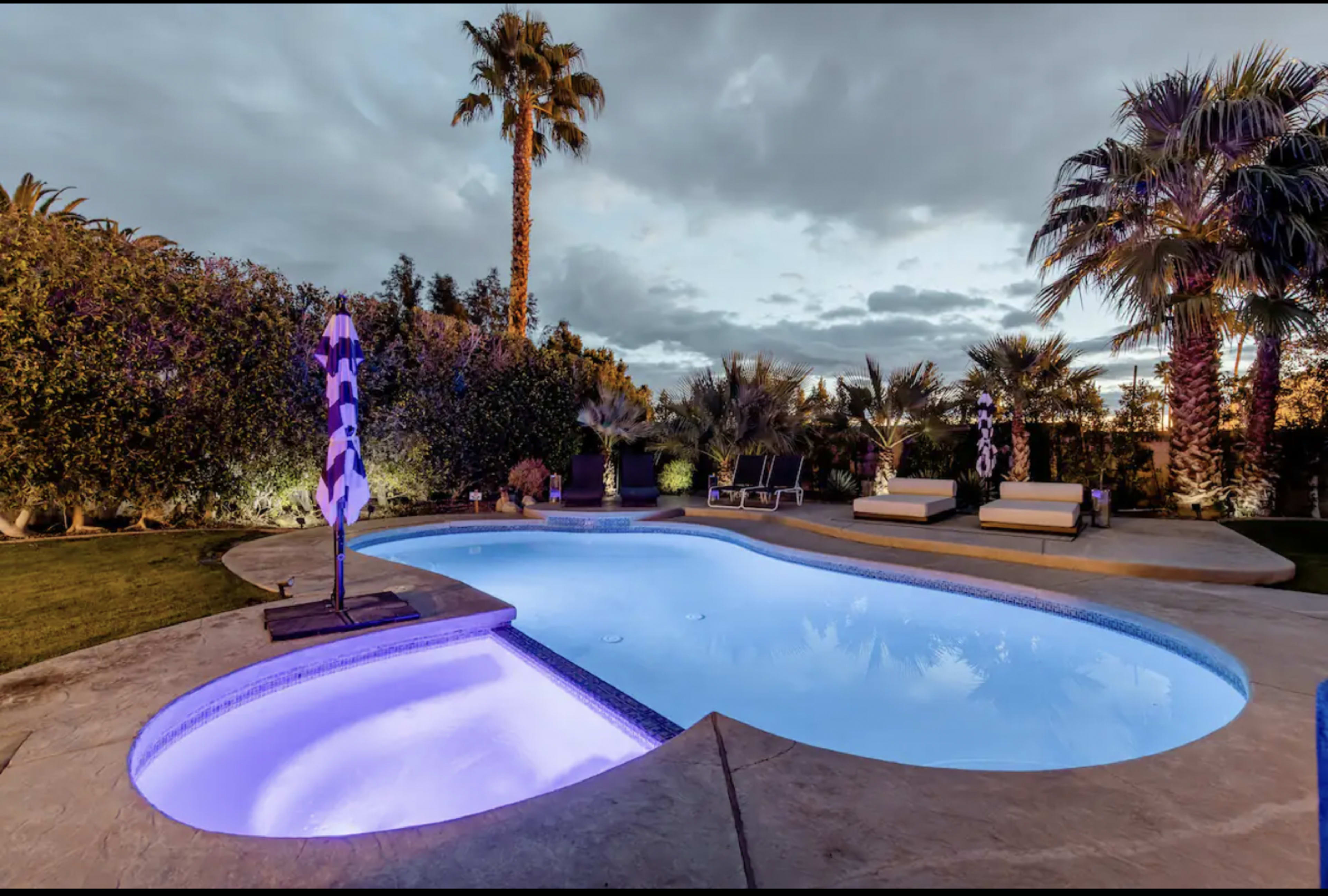 A swimming pool with illuminated water, surrounded by palm trees, lounge chairs, and a grassy area under a partly cloudy sky.