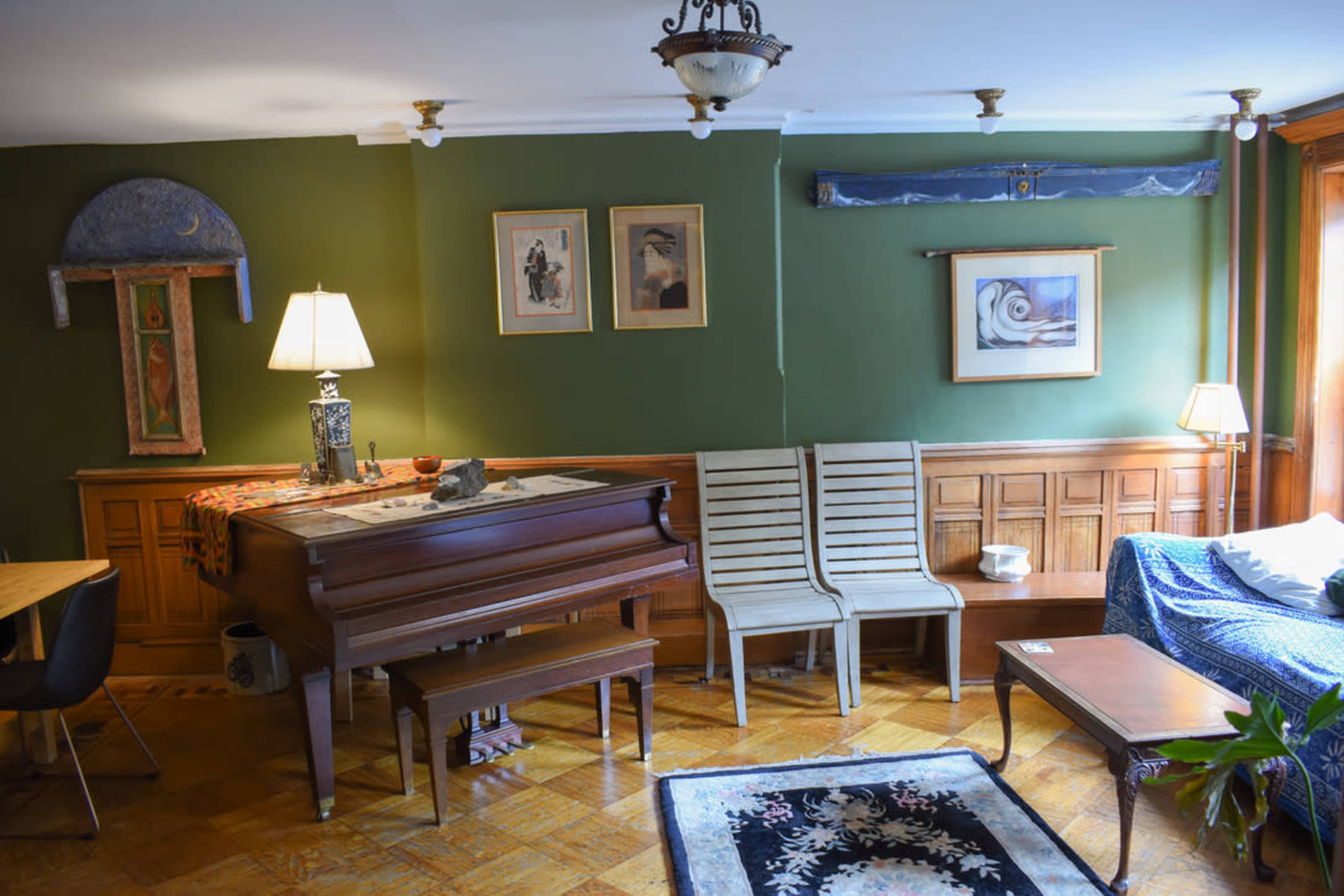 The room features a green wall with framed artwork, a piano, and a combination of wooden furniture, including chairs and a table.