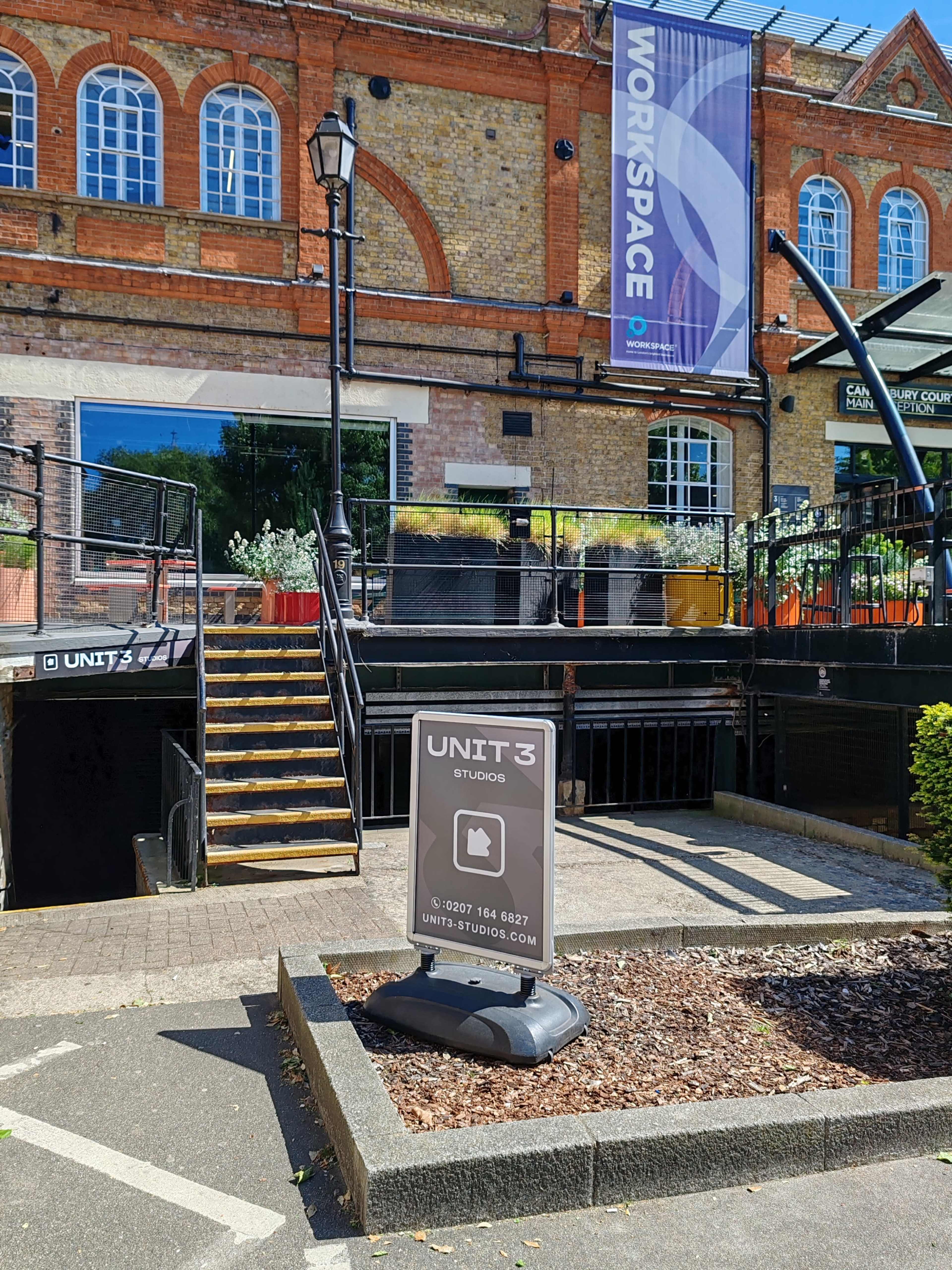 The image shows a sign for "Unit 3 Studios" in front of an entrance with stairs leading up to a building featuring a large banner.