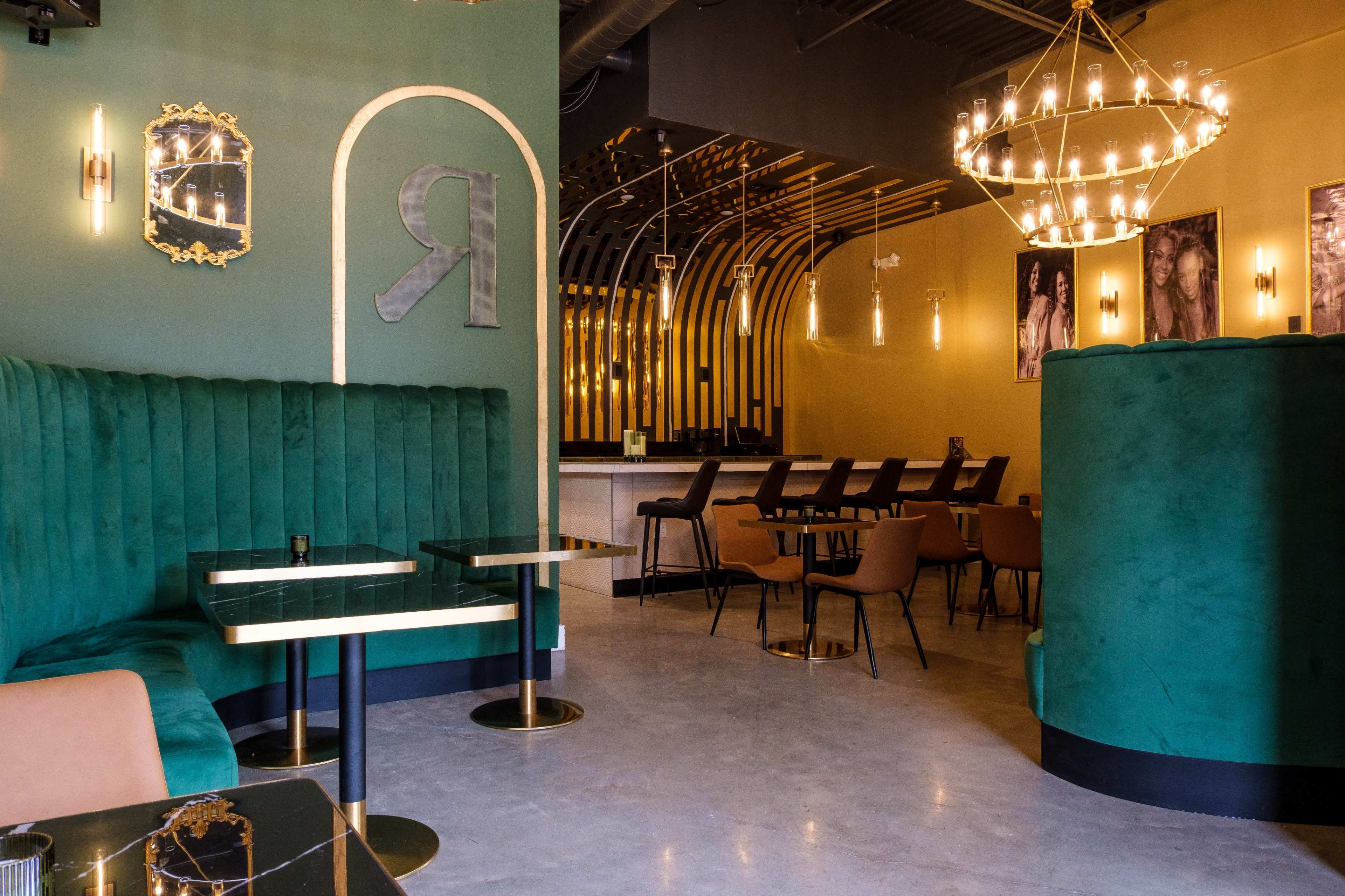 The image shows a modern café interior featuring plush green seating, elegant lighting fixtures, and a light-colored bar area in the background.