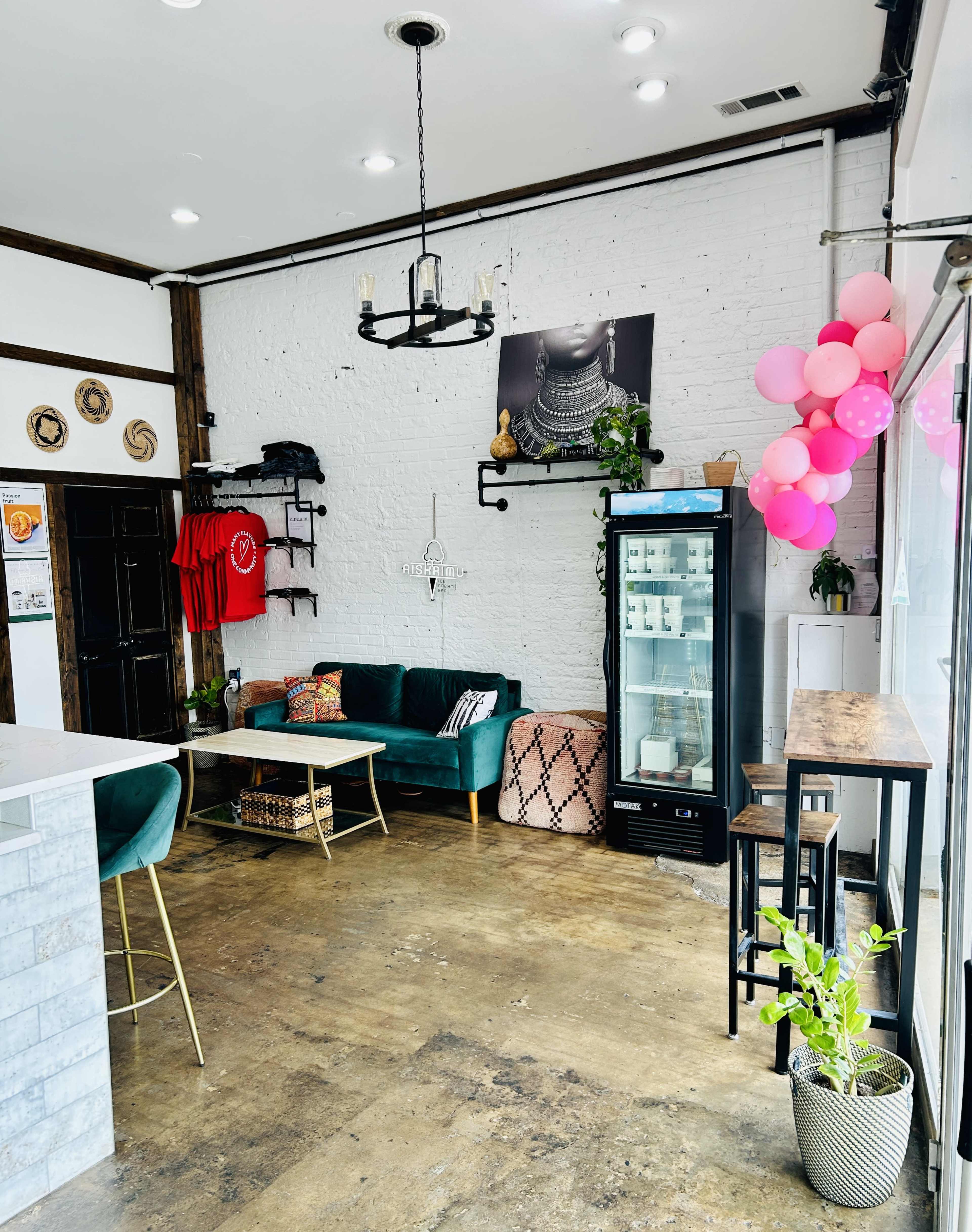 The image shows a stylish café interior featuring a green couch, a small table, and a beverage fridge against a white brick wall, adorned with colorful balloons and decorations.