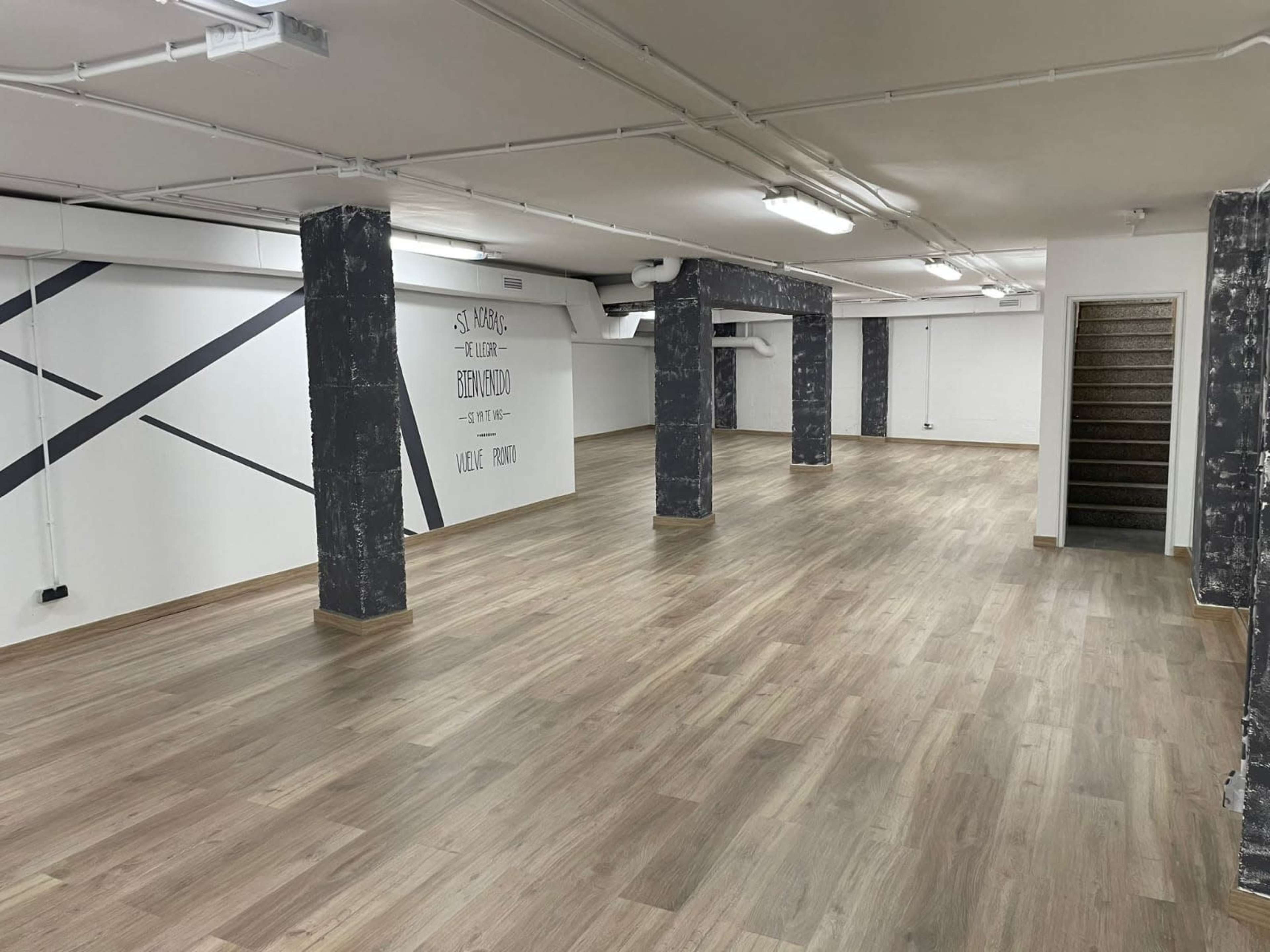 A spacious, empty indoor area with wooden floors, white walls featuring black geometric lines, and concrete columns, along with a set of stairs in the corner.