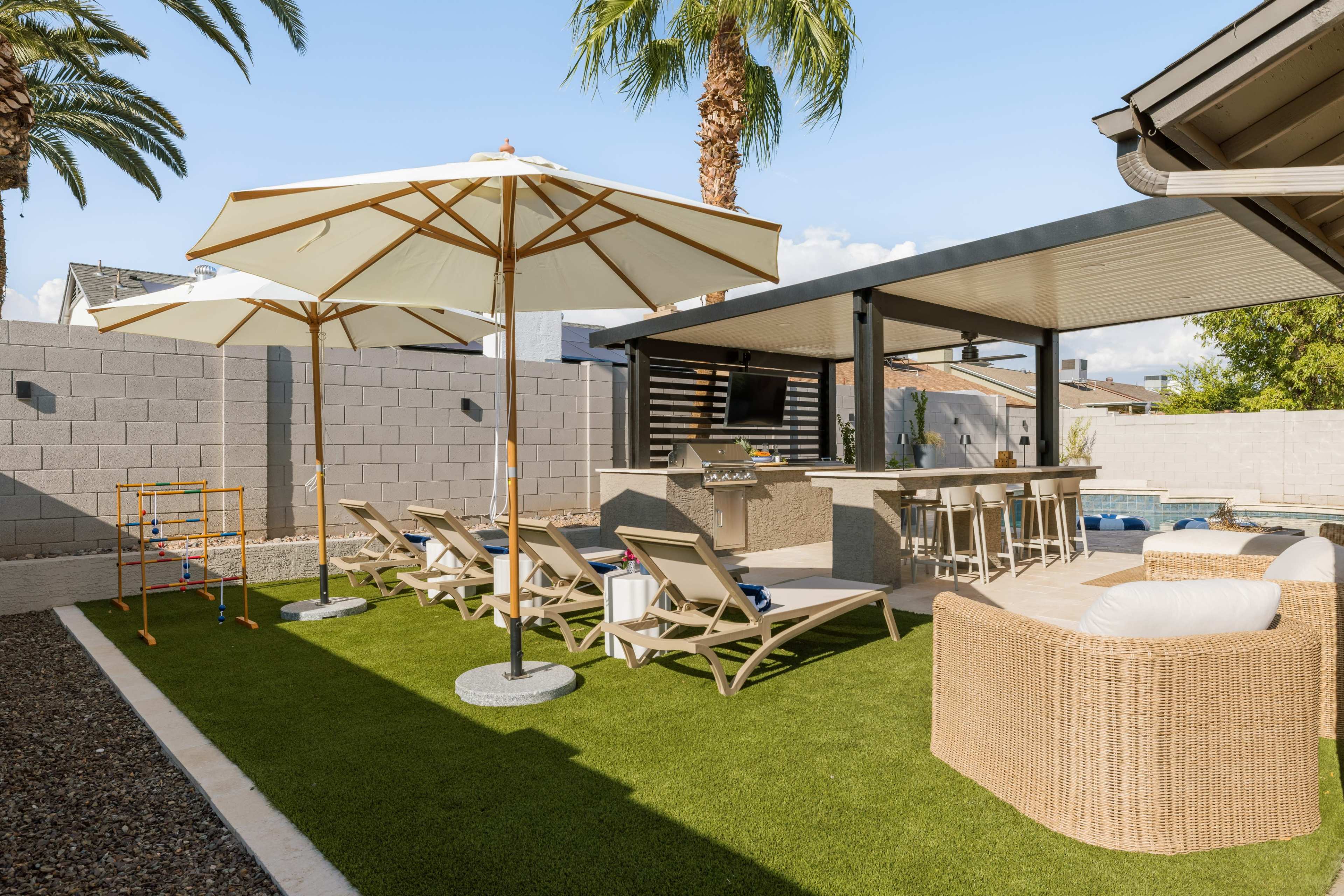 A spacious backyard features lounge chairs under large umbrellas, a shaded outdoor kitchen area, and palm trees against a clear blue sky.