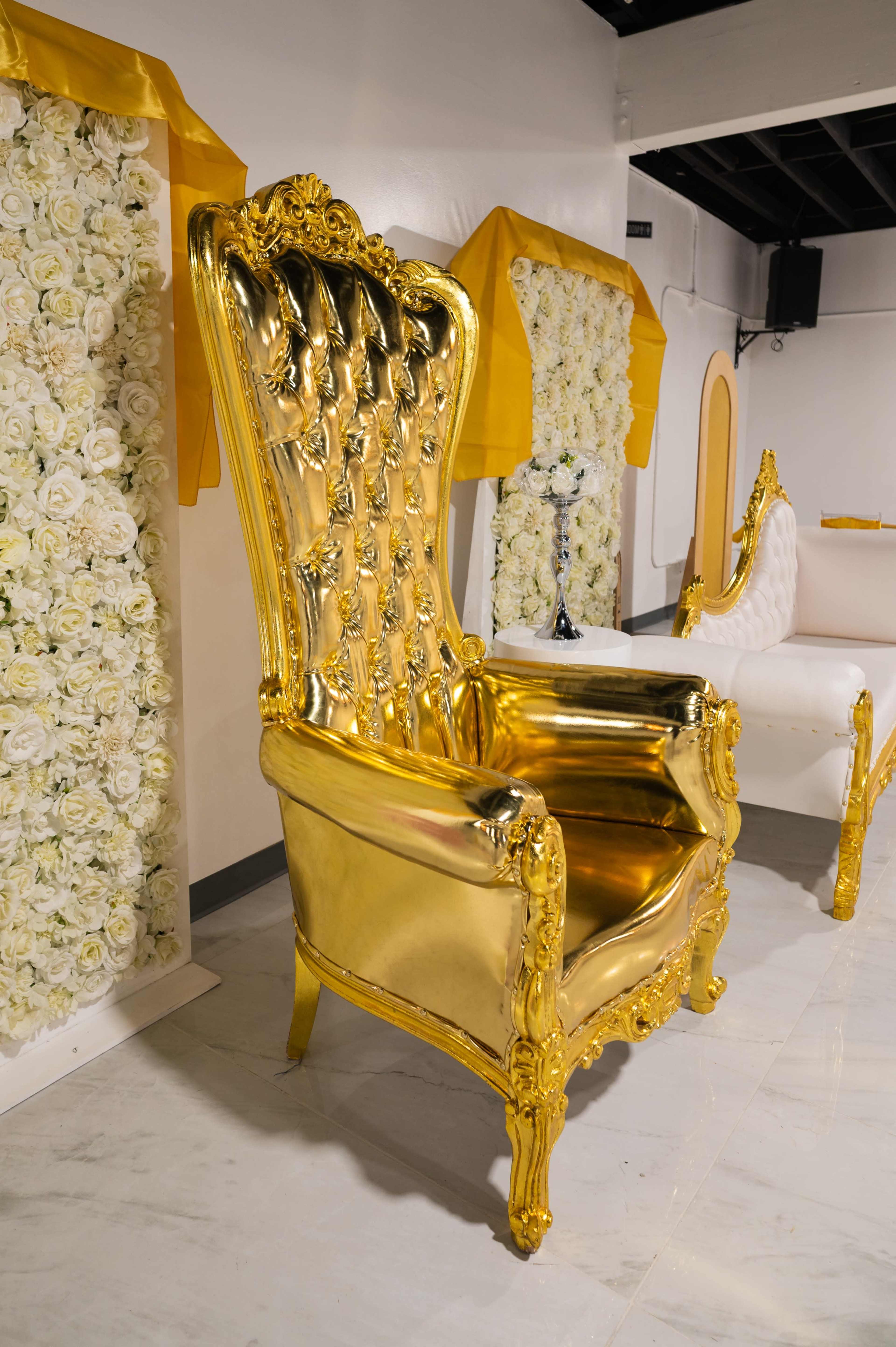 The image shows a large, ornate gold throne chair with a tufted back, situated in a room adorned with floral decorations.