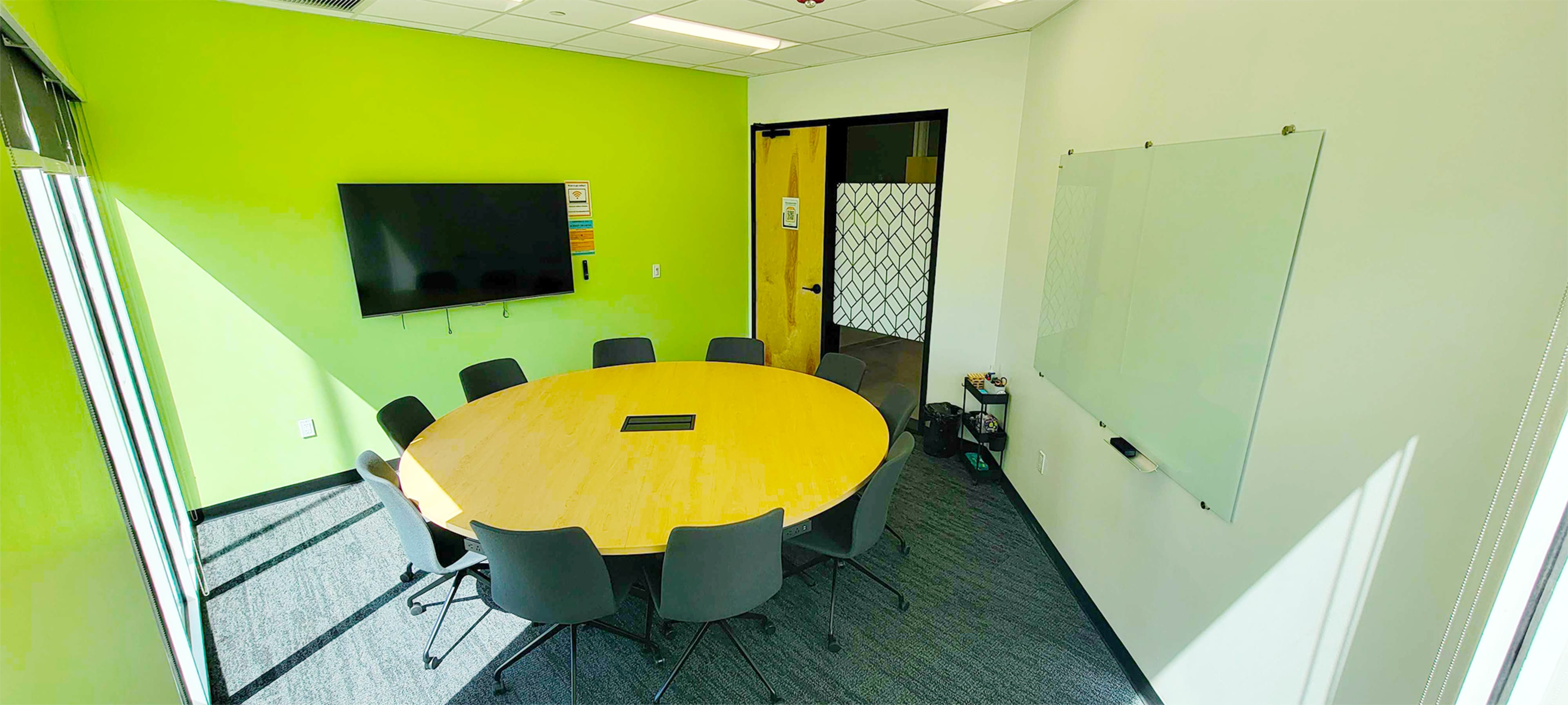 A modern conference room with a round wooden table surrounded by grey chairs, a wall-mounted television, and a whiteboard on one wall.