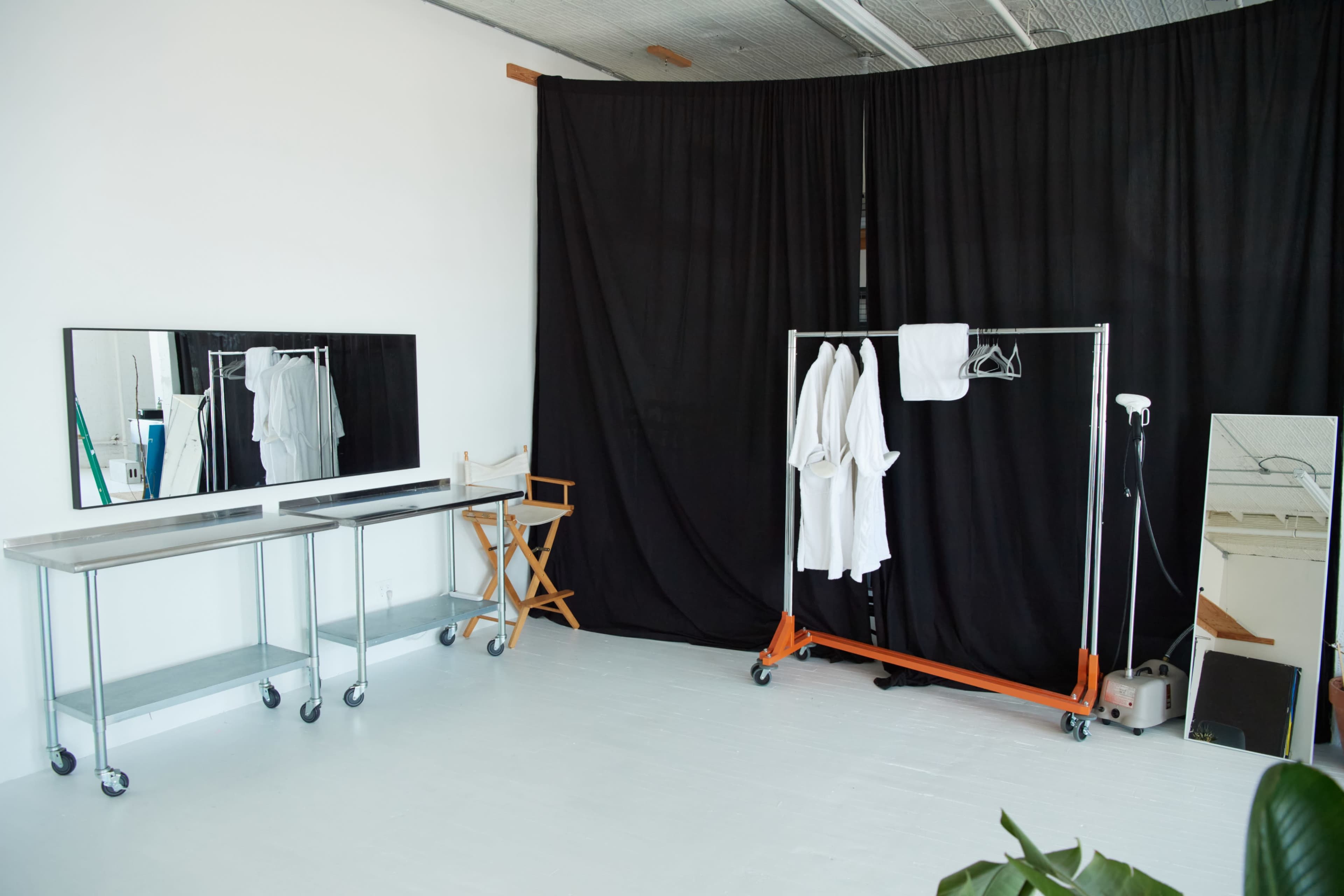 The image shows a simple studio space featuring a portable clothing rack with hanging garments, a mirror, a director's chair, and a black curtain dividing the area.