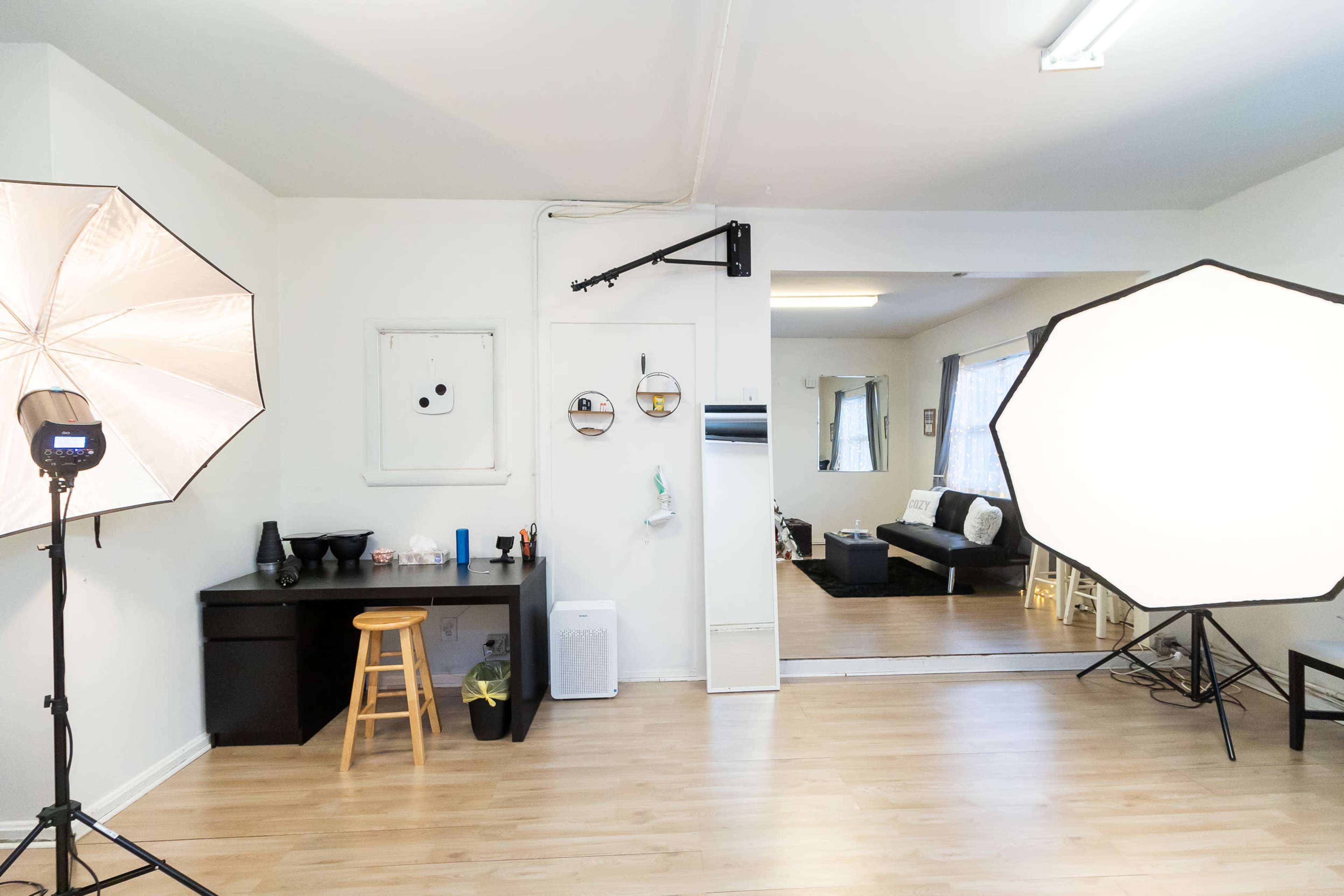 The image shows a bright room with a small desk and stool on one side, two large softbox lights positioned for a photo setup, and a living area with a sofa in the background.