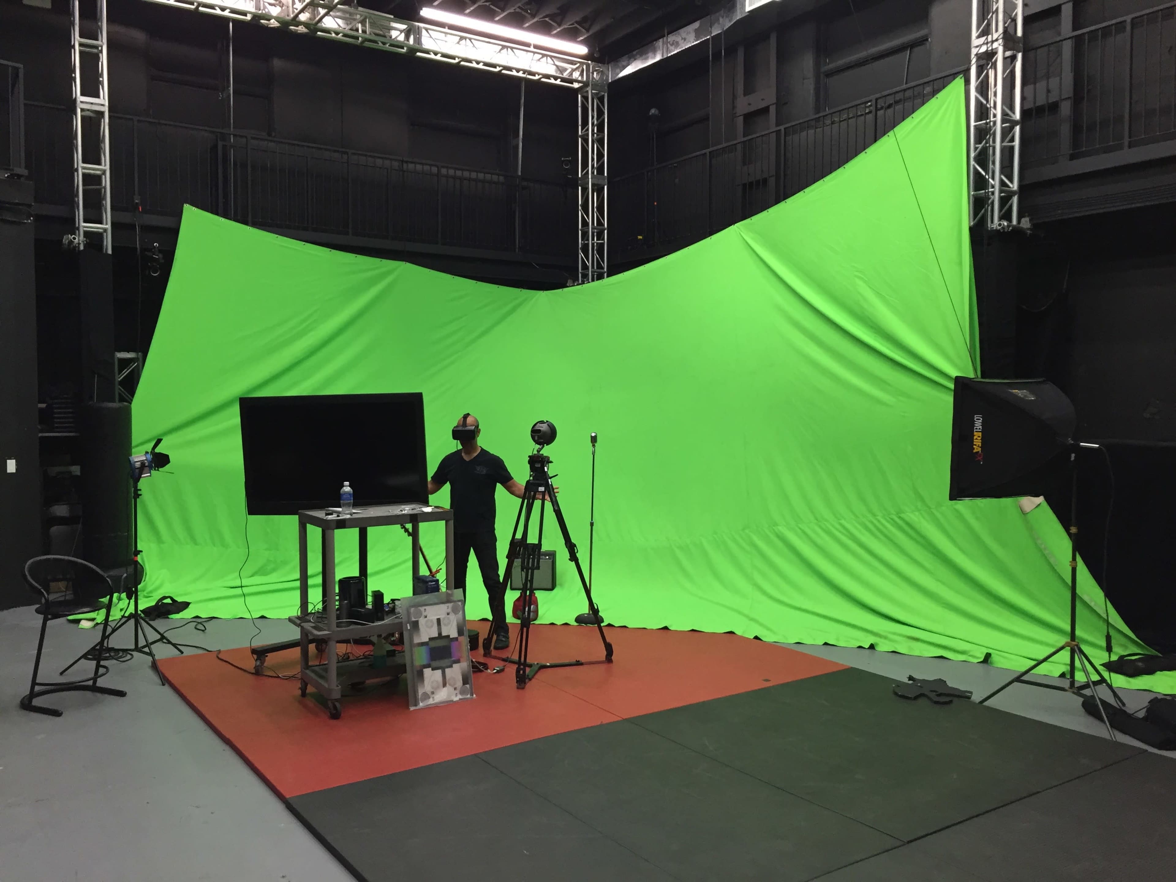 A green screen studio setup features a large green backdrop, a table with equipment, cameras, and lighting in an indoor space.