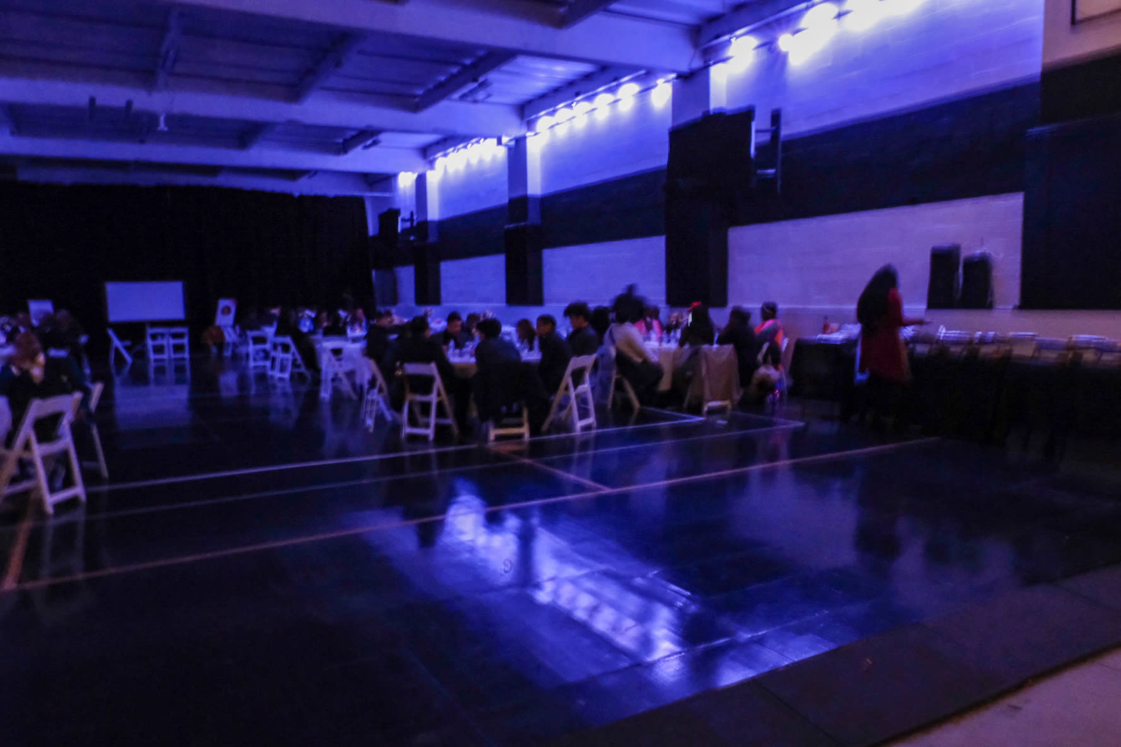 A large gymnasium is dimly lit in blue light, with rows of tables set for an event and people seated throughout the space.