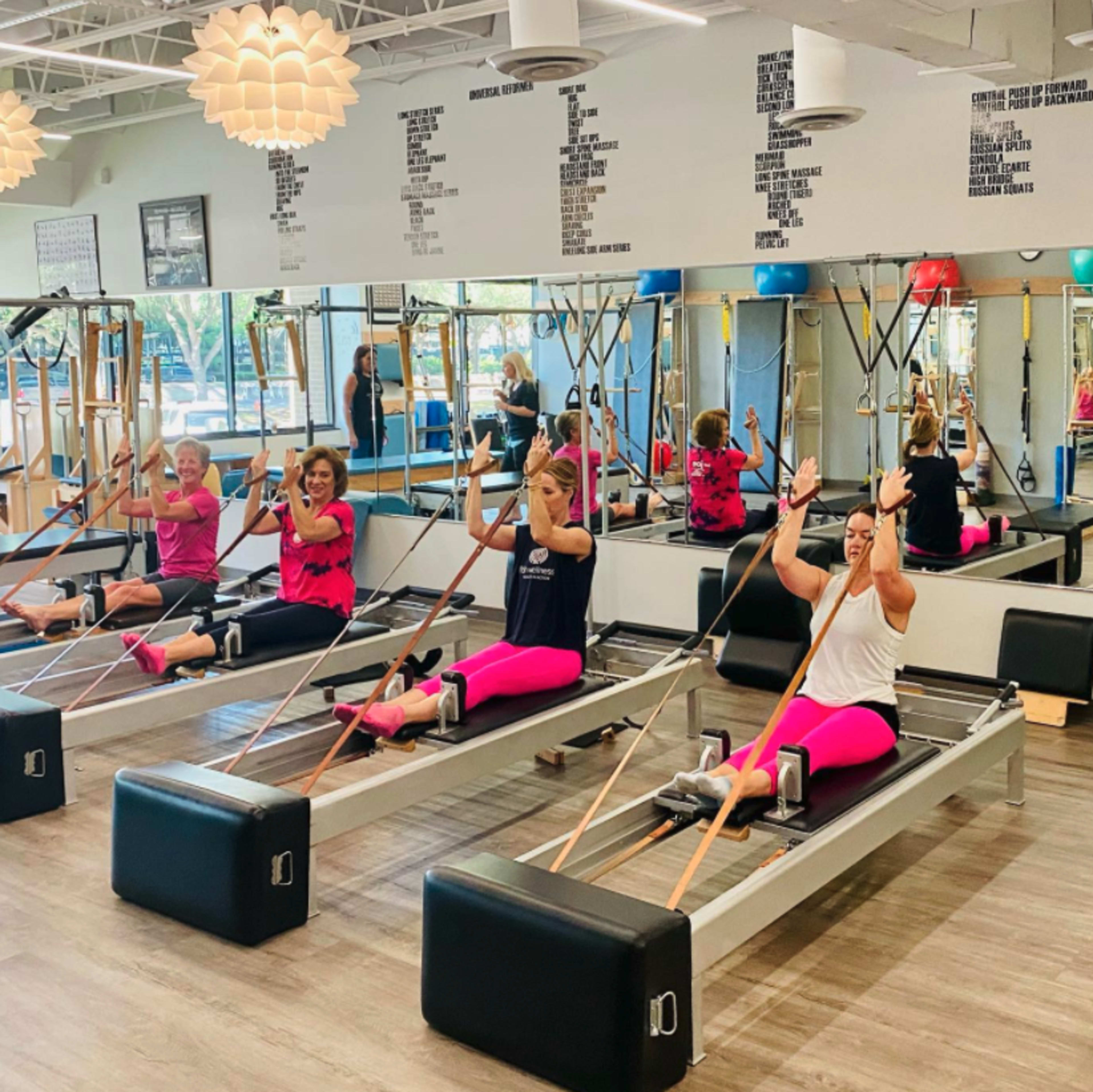 A group of five women are performing an exercise class on reformer machines in a brightly lit studio.