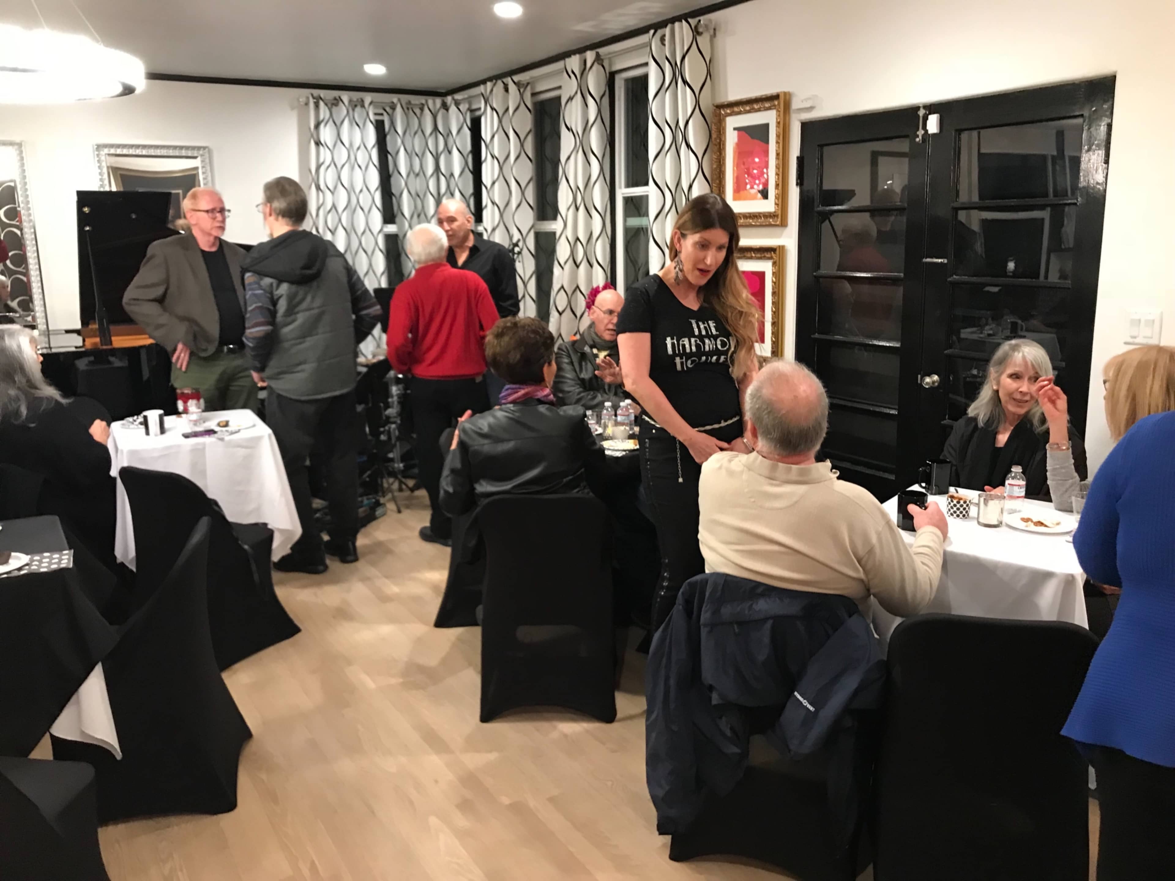 A gathering of people socializes in a well-decorated room with tables and a piano in the background.