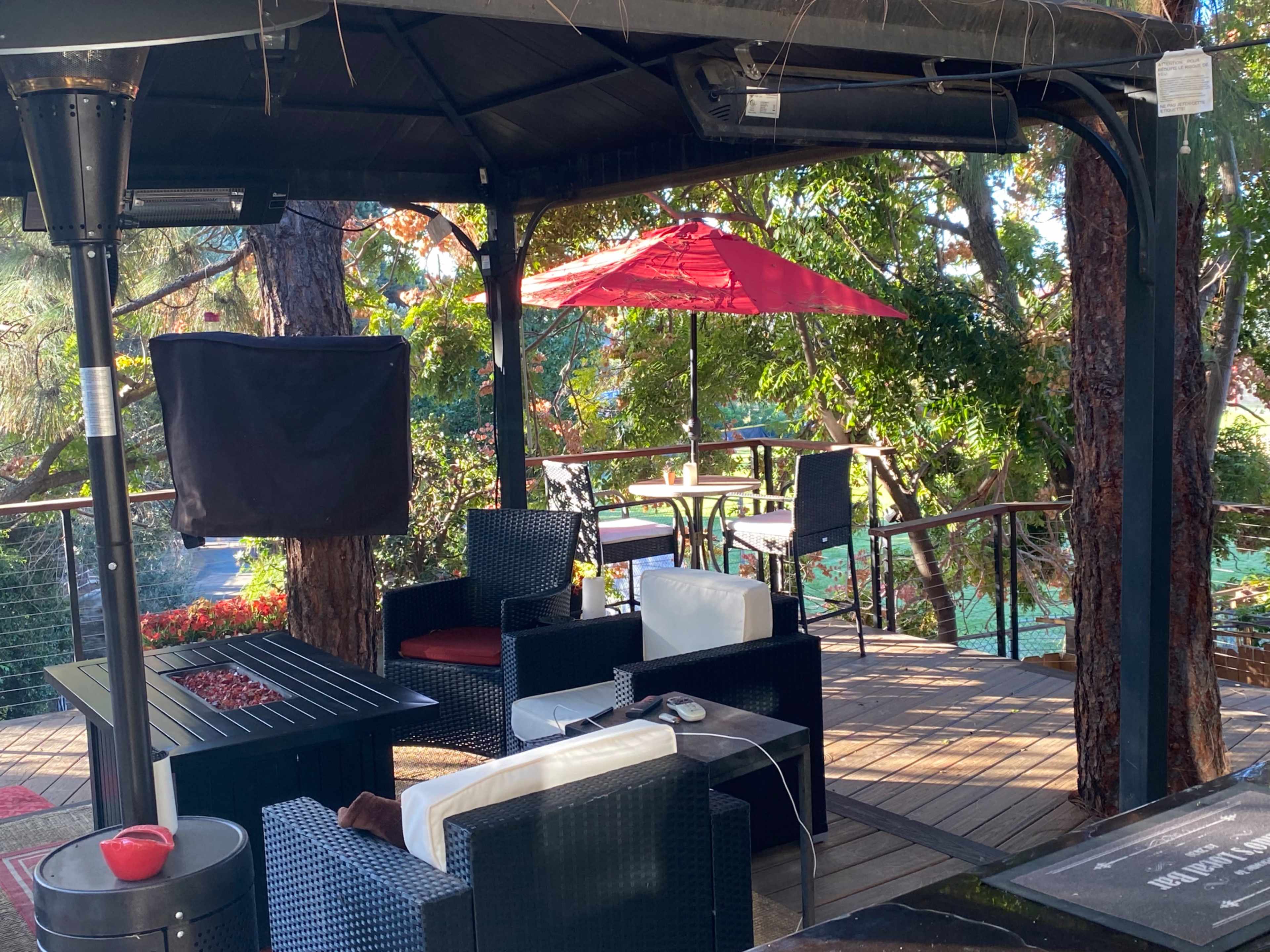 A covered outdoor seating area with black wicker furniture and a red umbrella on a wooden deck surrounded by trees.