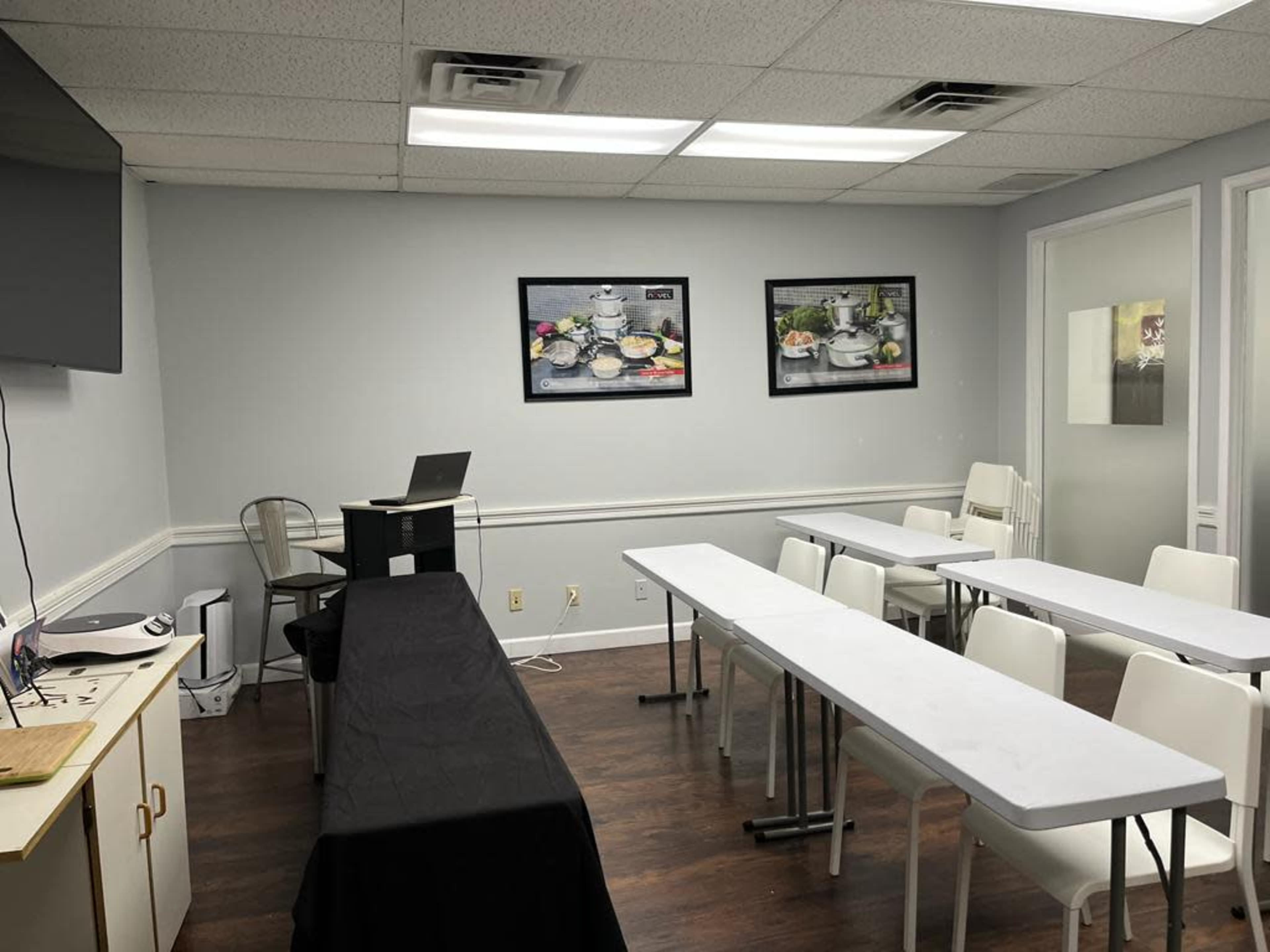 A small, well-lit meeting room features a black table at the front, rows of white tables and chairs, and two framed pictures on the walls.