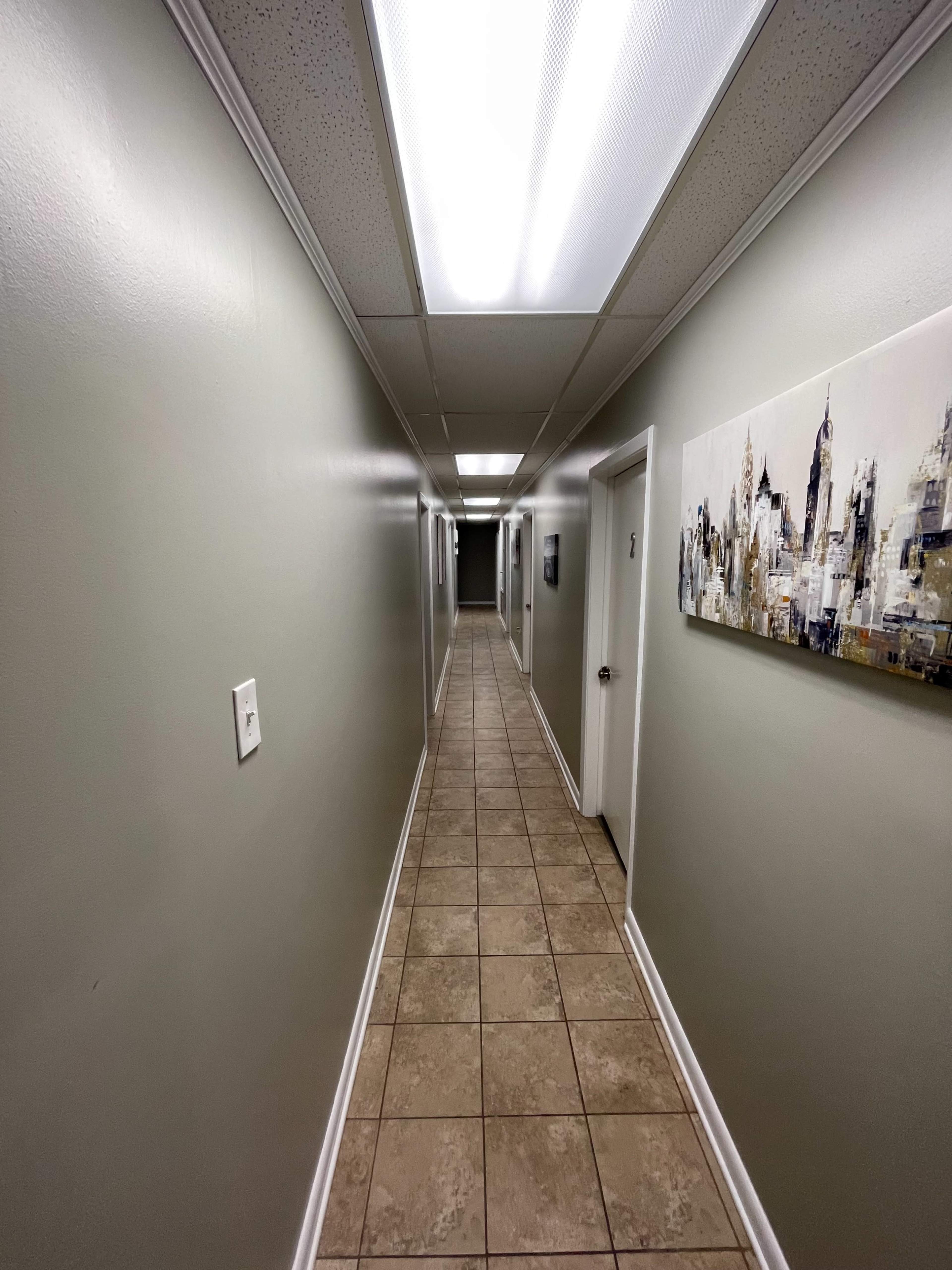 A long, narrow hallway with light-colored walls, tile flooring, and several closed doors on either side.