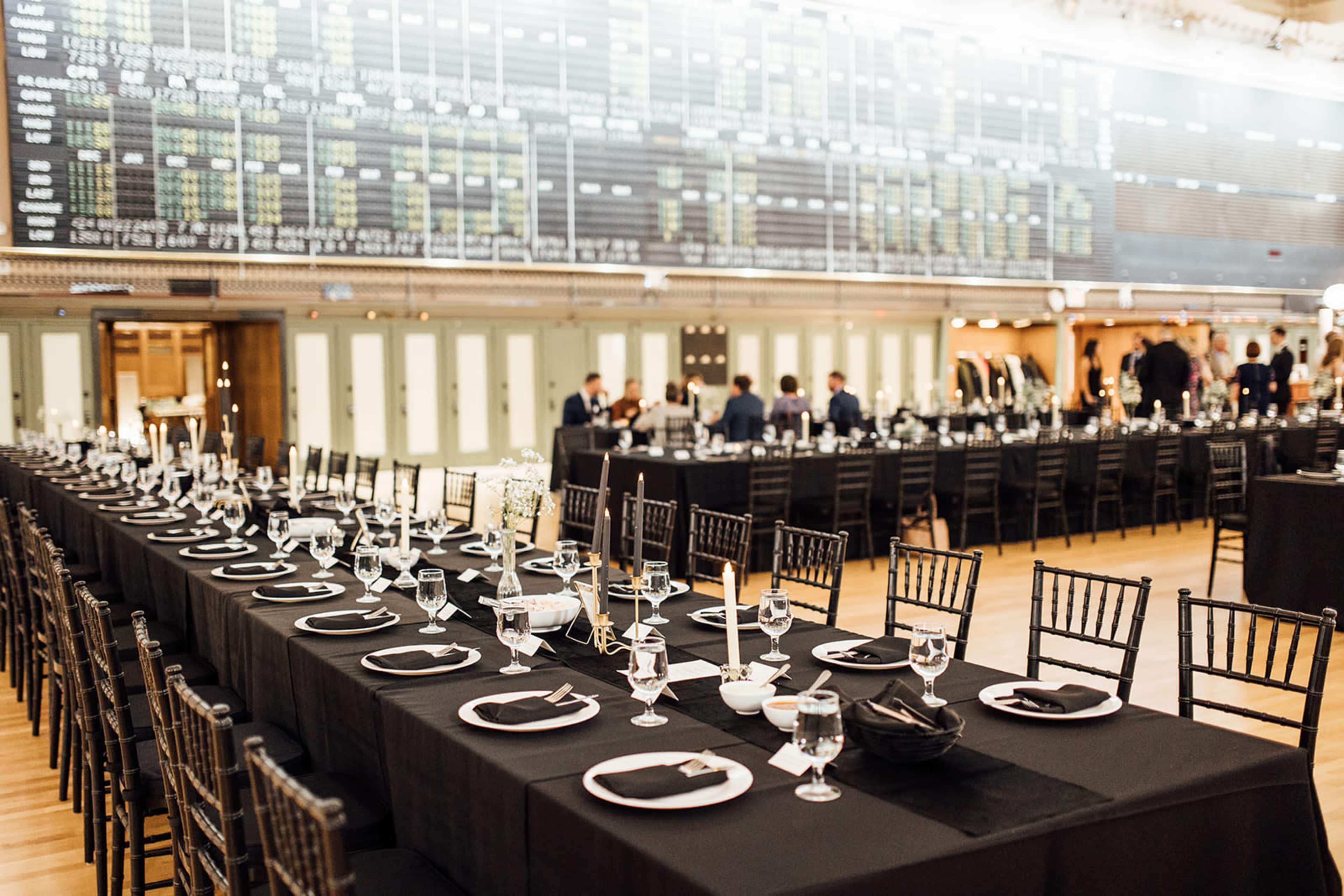 A long dining arrangement is set with black tablecloths, plates, and glassware in a spacious room featuring a large display board in the background.