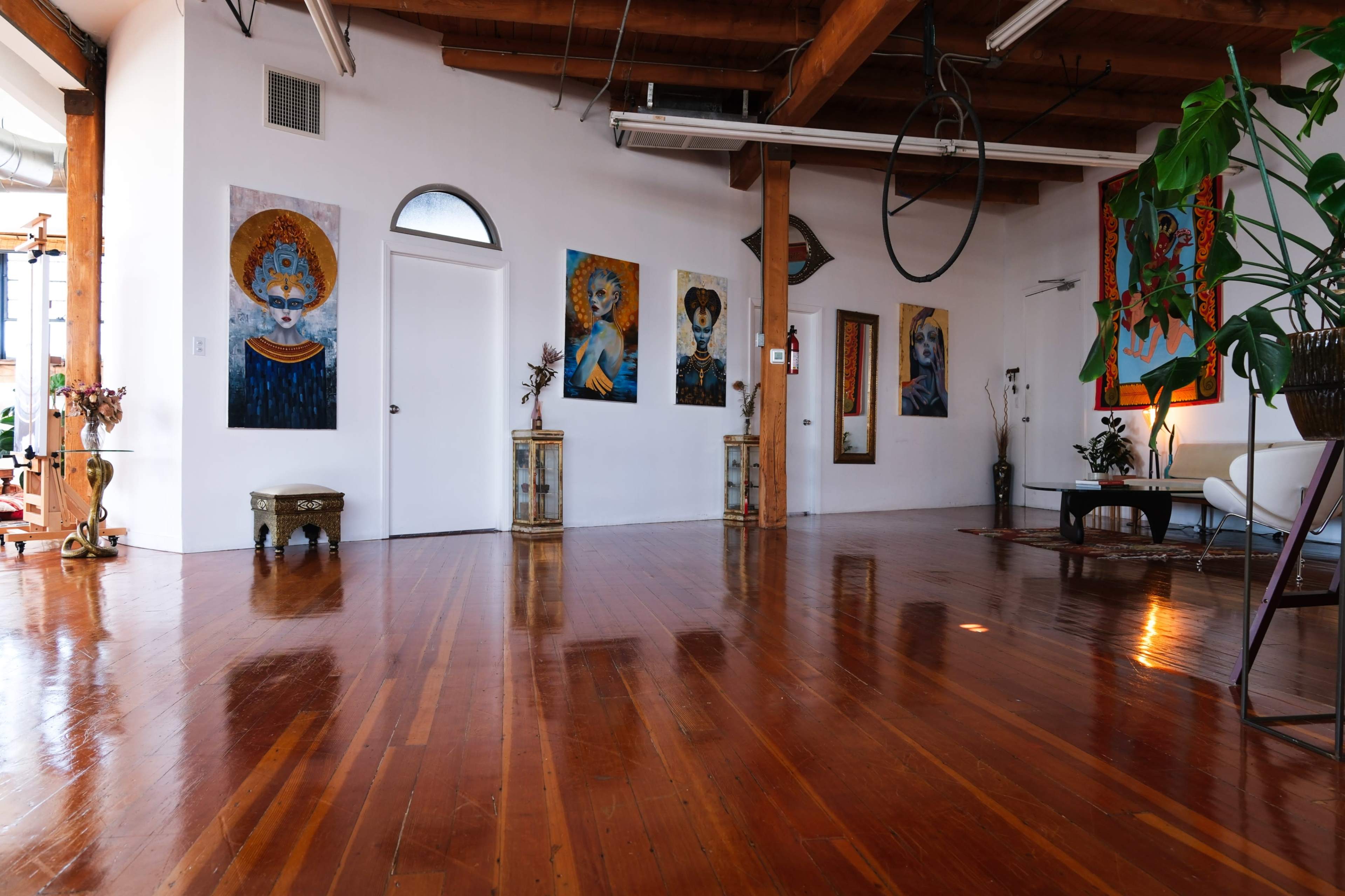 The image shows a spacious, well-lit room with polished wooden floors, featuring colorful paintings on the walls and modern furniture.