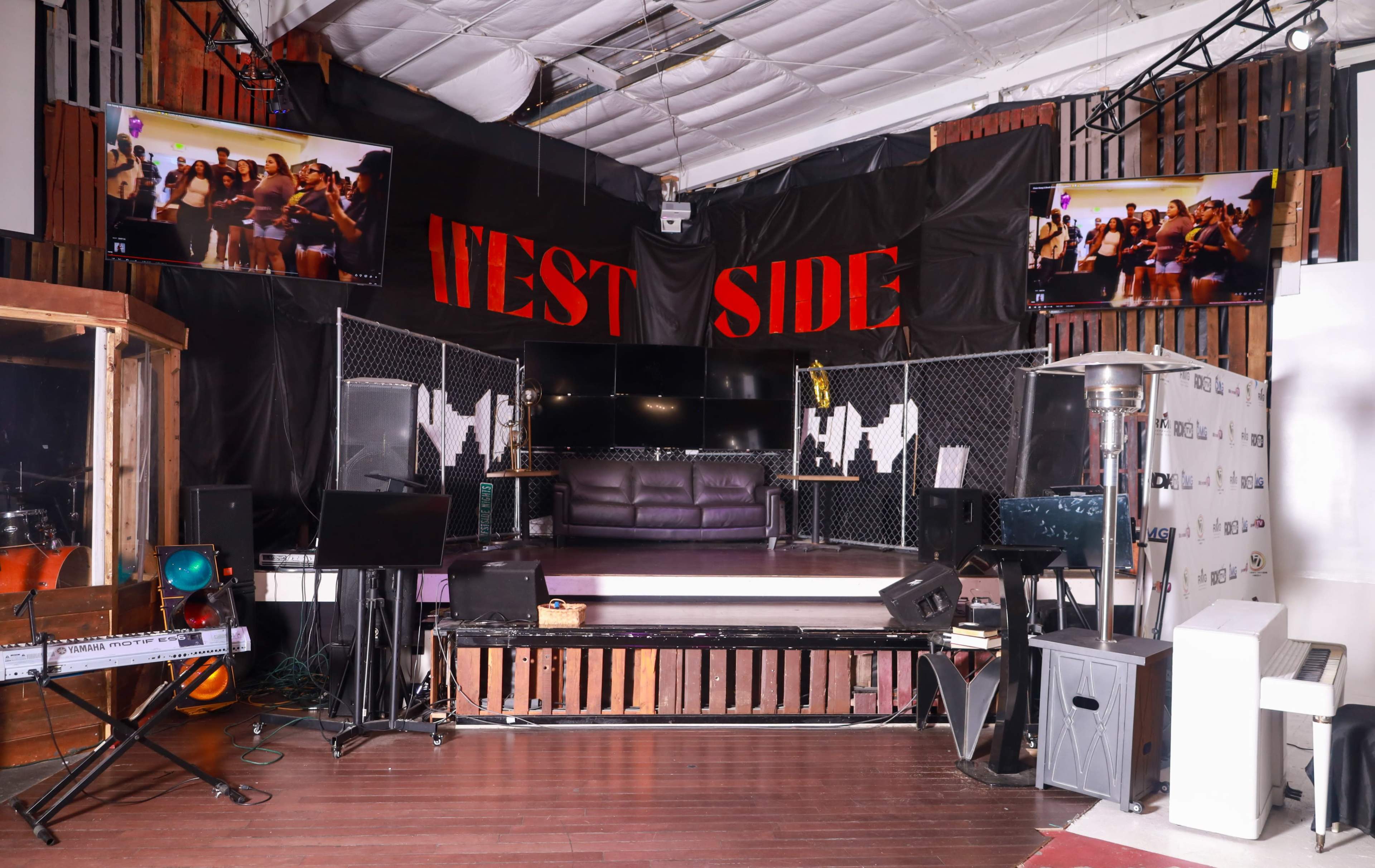 The image shows an indoor stage area with a black backdrop displaying "WEST SIDE," two video screens mounted on the walls, and various musical equipment arranged in front.